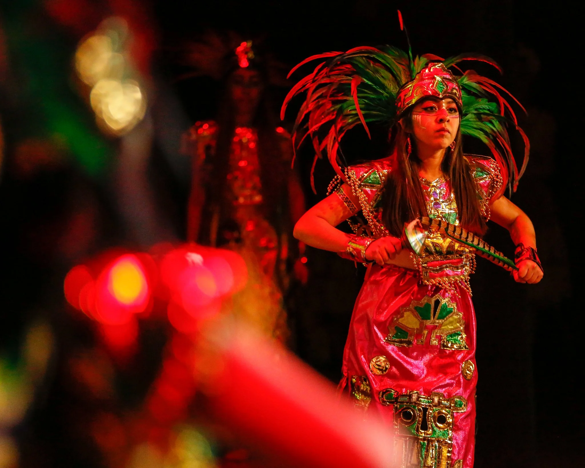 A young girl dressed in a colorful traditional costume with feathers, holding a stick, performing in a cultural dance or ceremony at night.