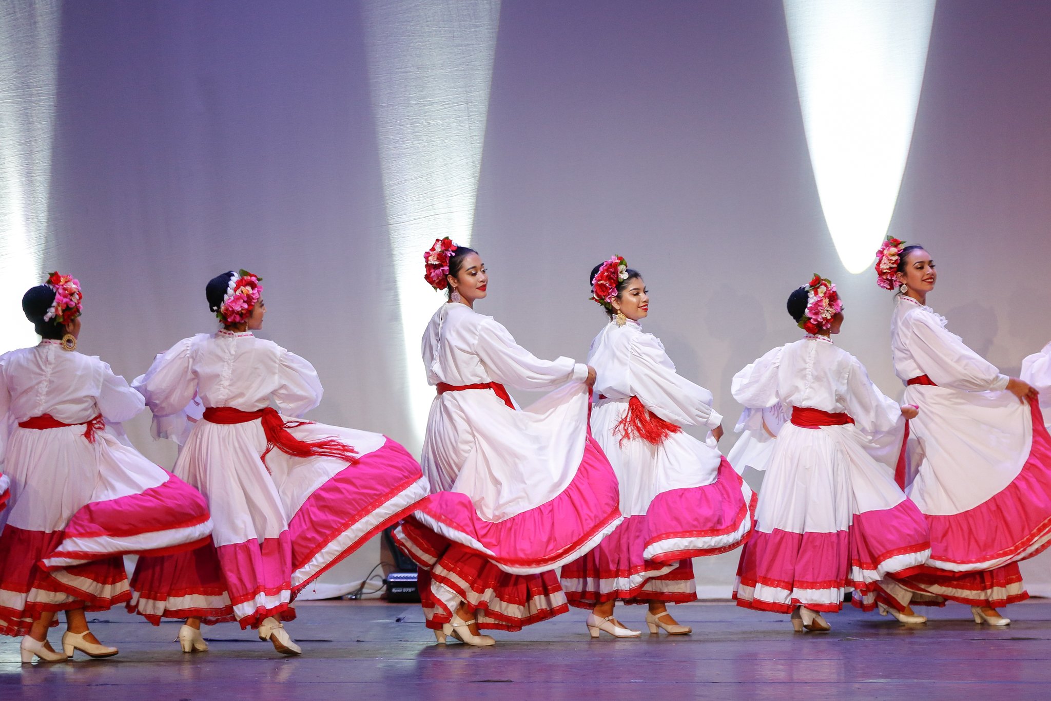 A group of women in traditional Mexican dance costumes, wearing white blouses, pink and red skirts, and floral headpieces, performing a dance on stage.