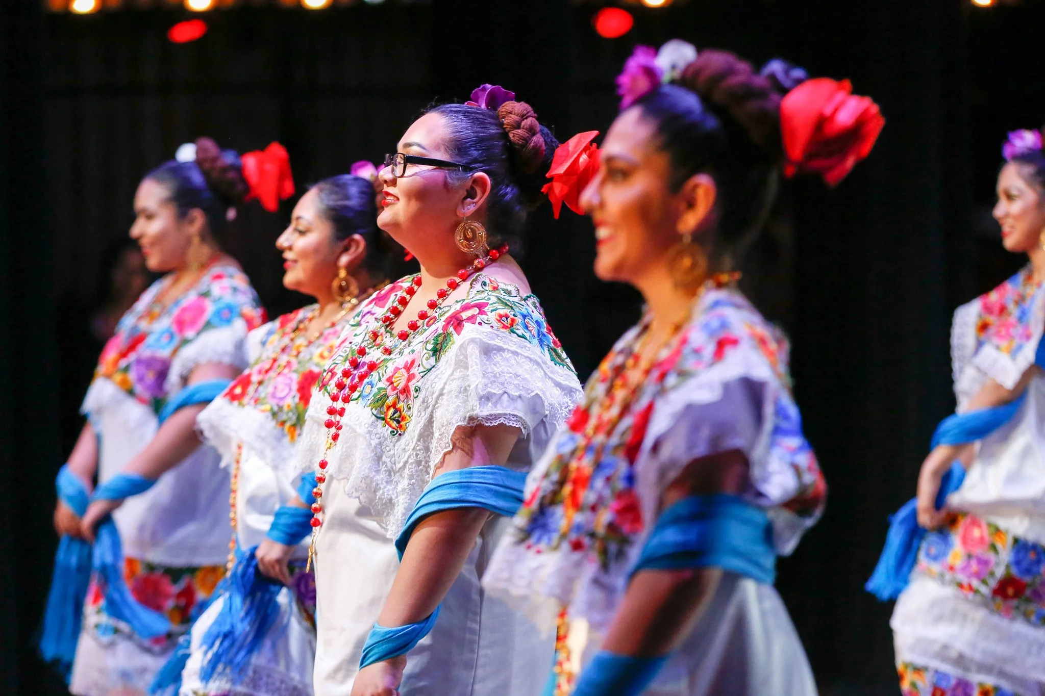 A group of women in traditional Mexican folk dance costumes with floral embroidery, colorful beads, and flowers in their hair, standing in a line and smiling.