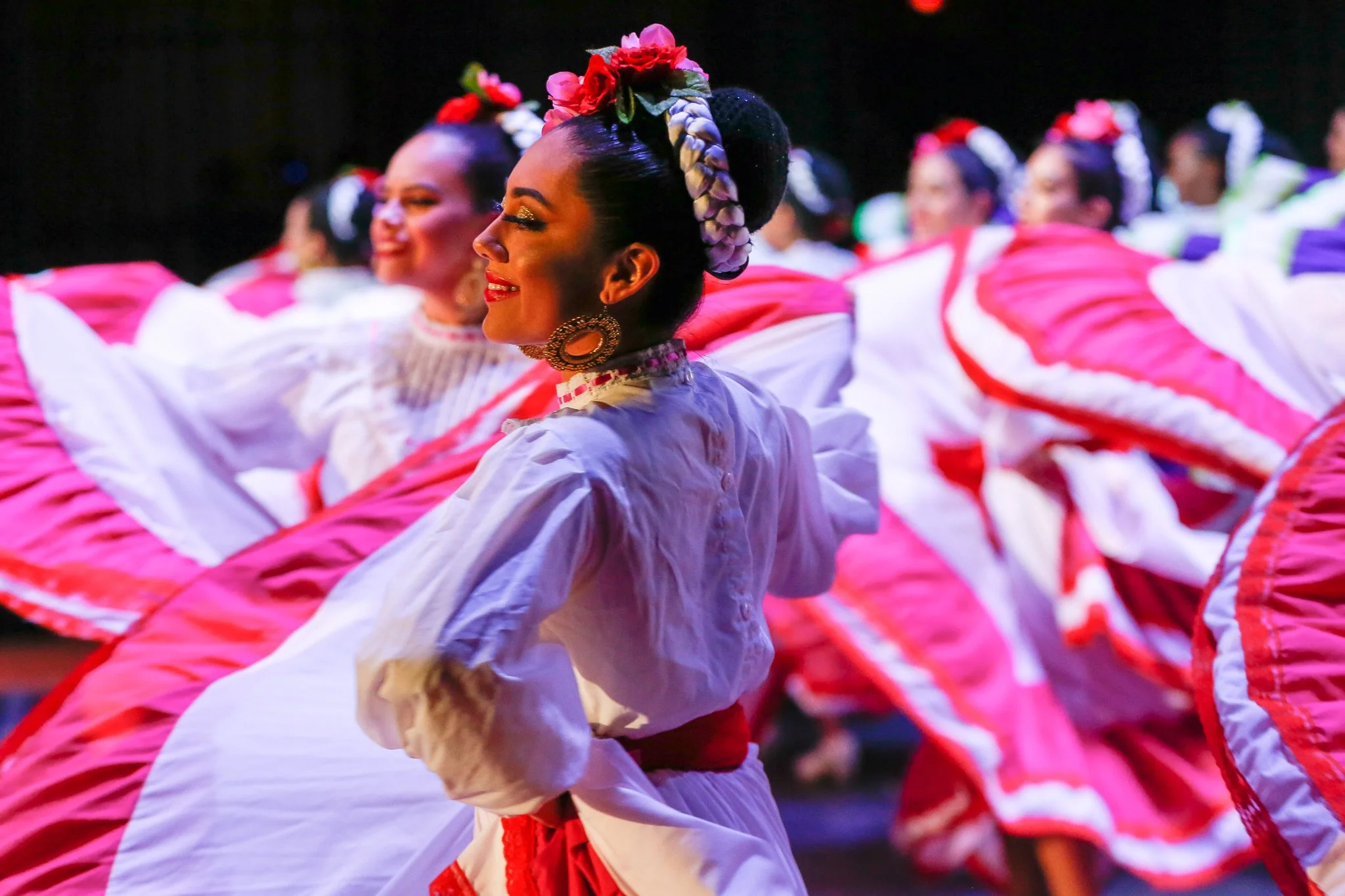 Women in colorful traditional Mexican dance costumes, smiling and performing a folkloric dance with pink and white skirts and floral headpieces.