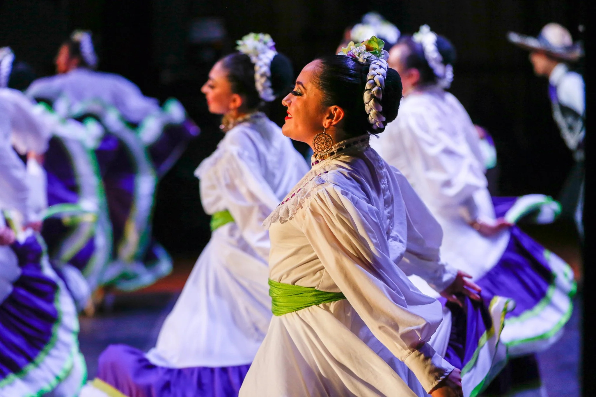 Women dressed in traditional Mexican folkloric attire performing a dance on stage, with dark background and colorful costumes.