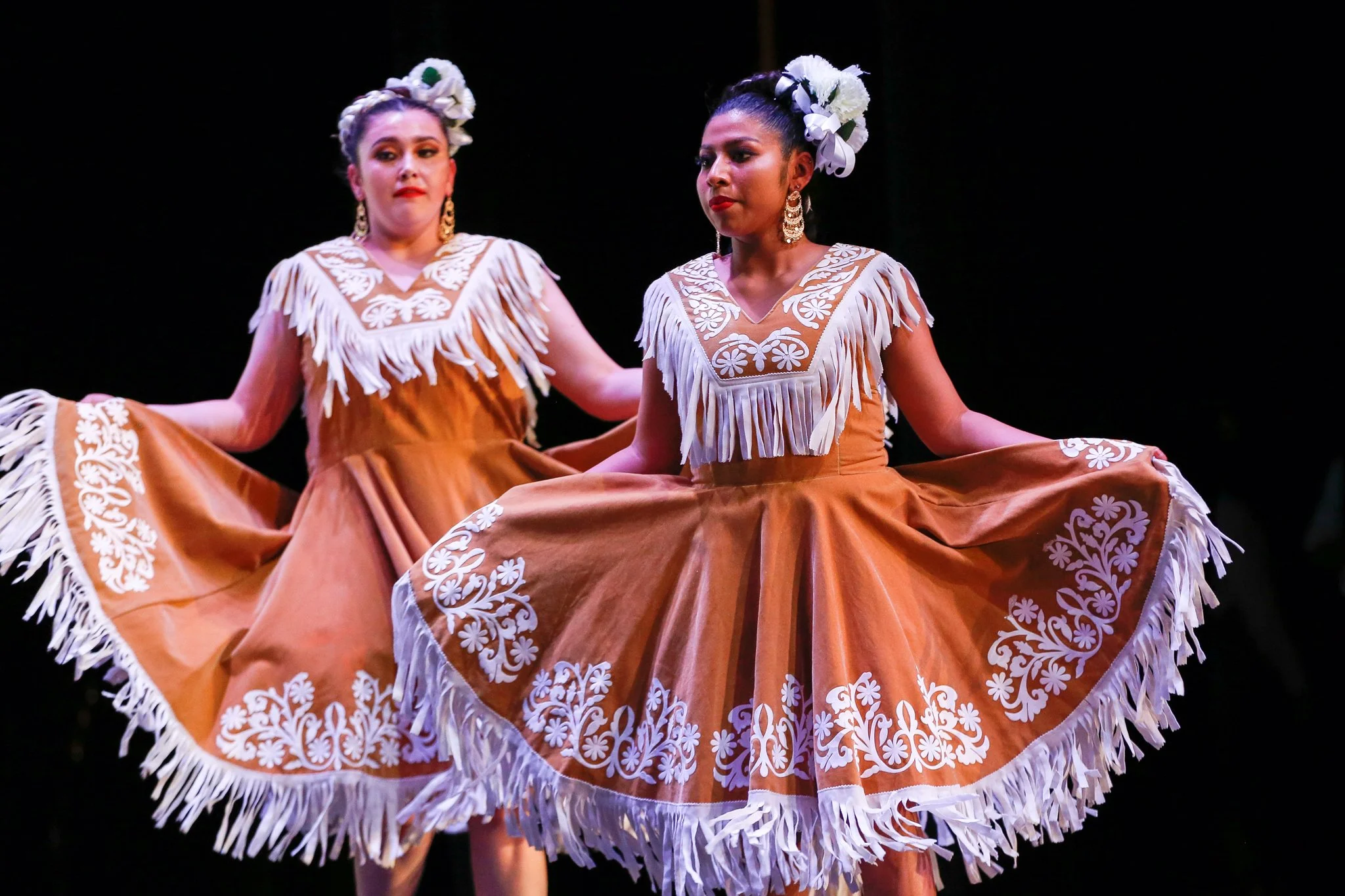 Two women dressed in traditional Mexican folkloric dresses with white floral embroidery and fringe, performing a cultural dance on stage.
