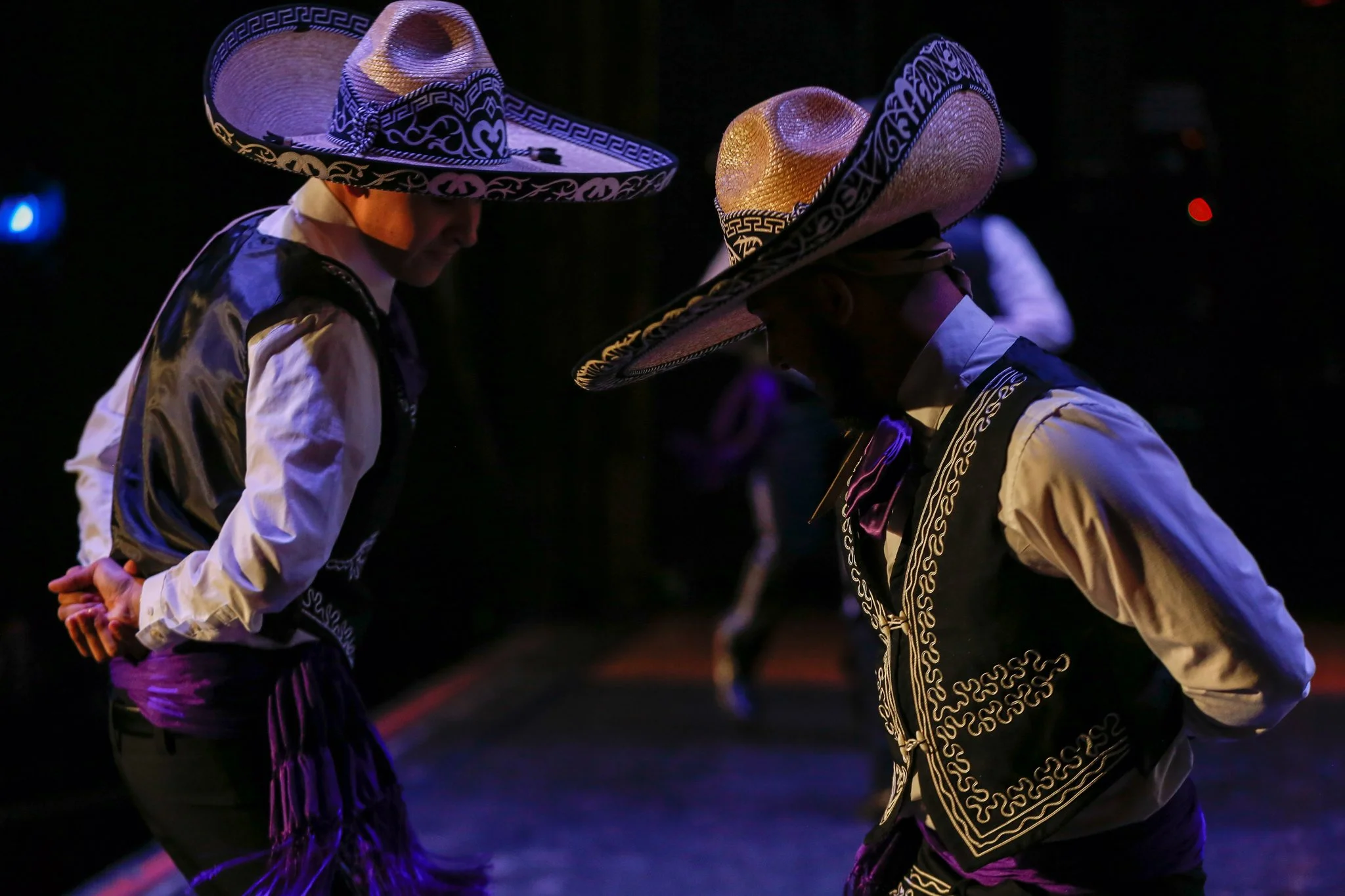 Two people dressed in traditional Mexican charro and mariachi attire, wearing large sombreros and bow ties, are bowing to each other in a dark setting with colorful lighting.