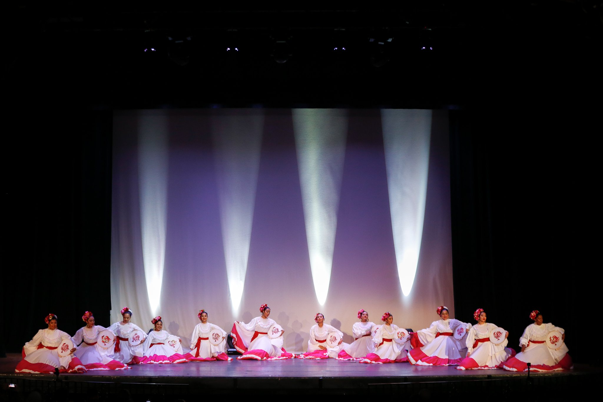 Group of dancers in traditional white and red dresses performing on stage with spotlight effects.