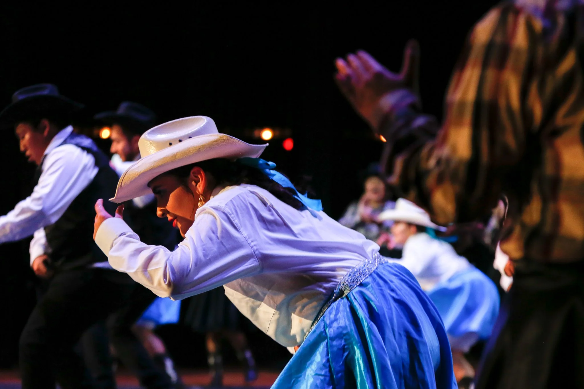 People dancing in traditional cowboy attire, including hats, on a dark stage.