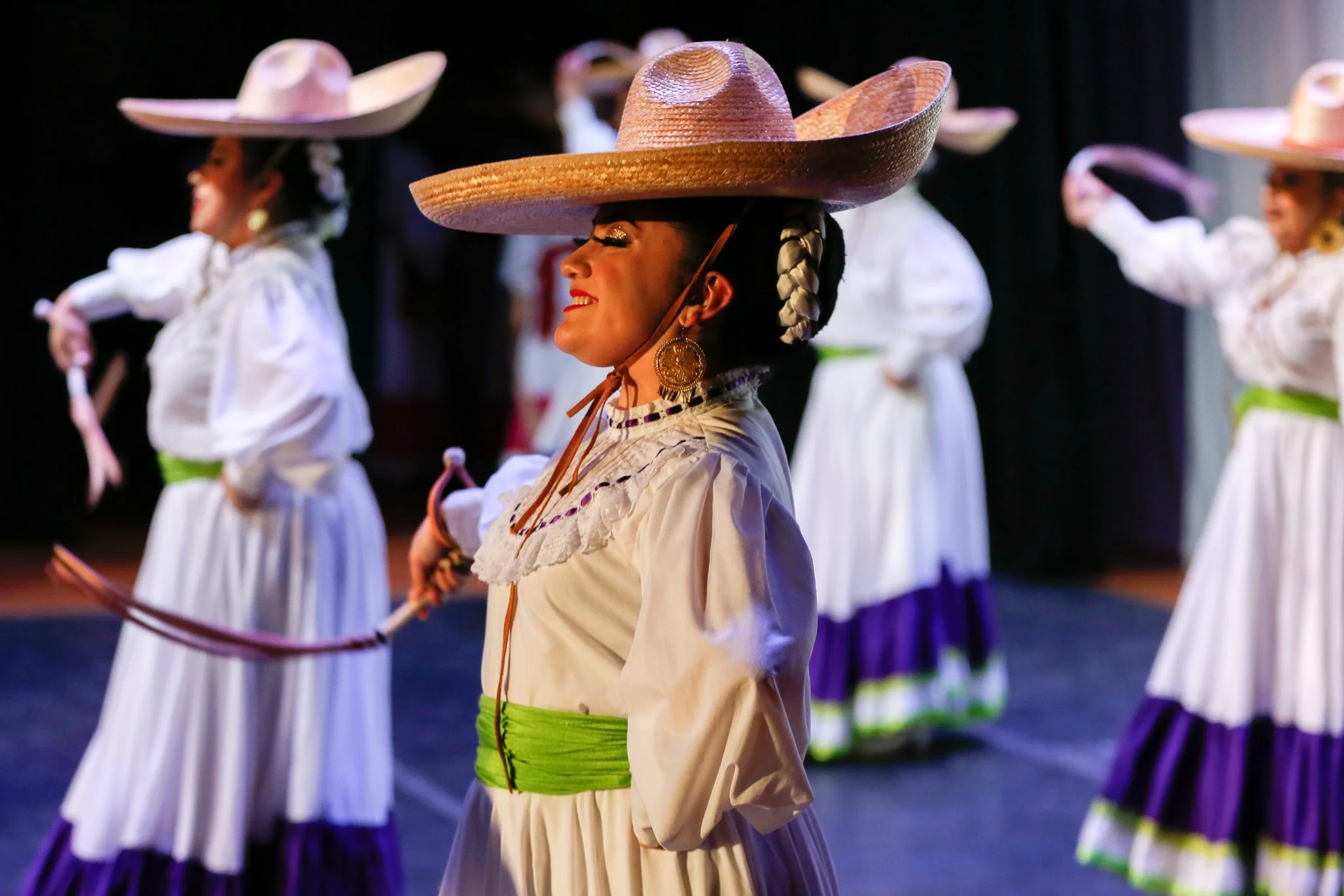 Group of women dressed in traditional Mexican folkloric attire performing a dance, wearing colorful wide-brimmed sombreros, white blouses, and long skirts with purple and green accents.