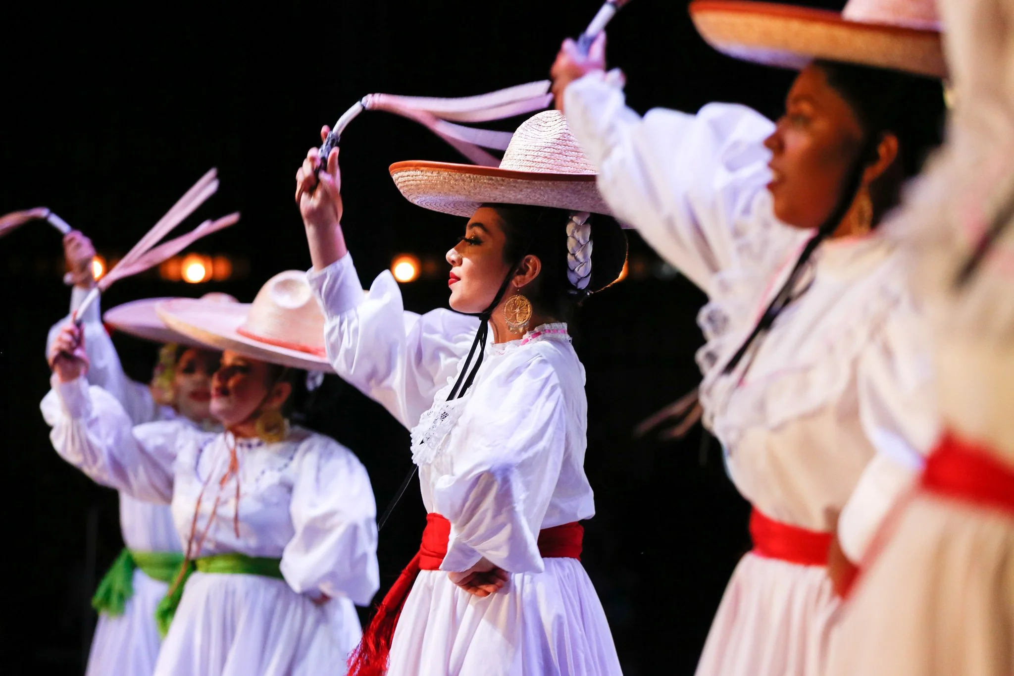 Women dressed in traditional white dresses with colorful sashes and large straw hats, performing a dance at night.