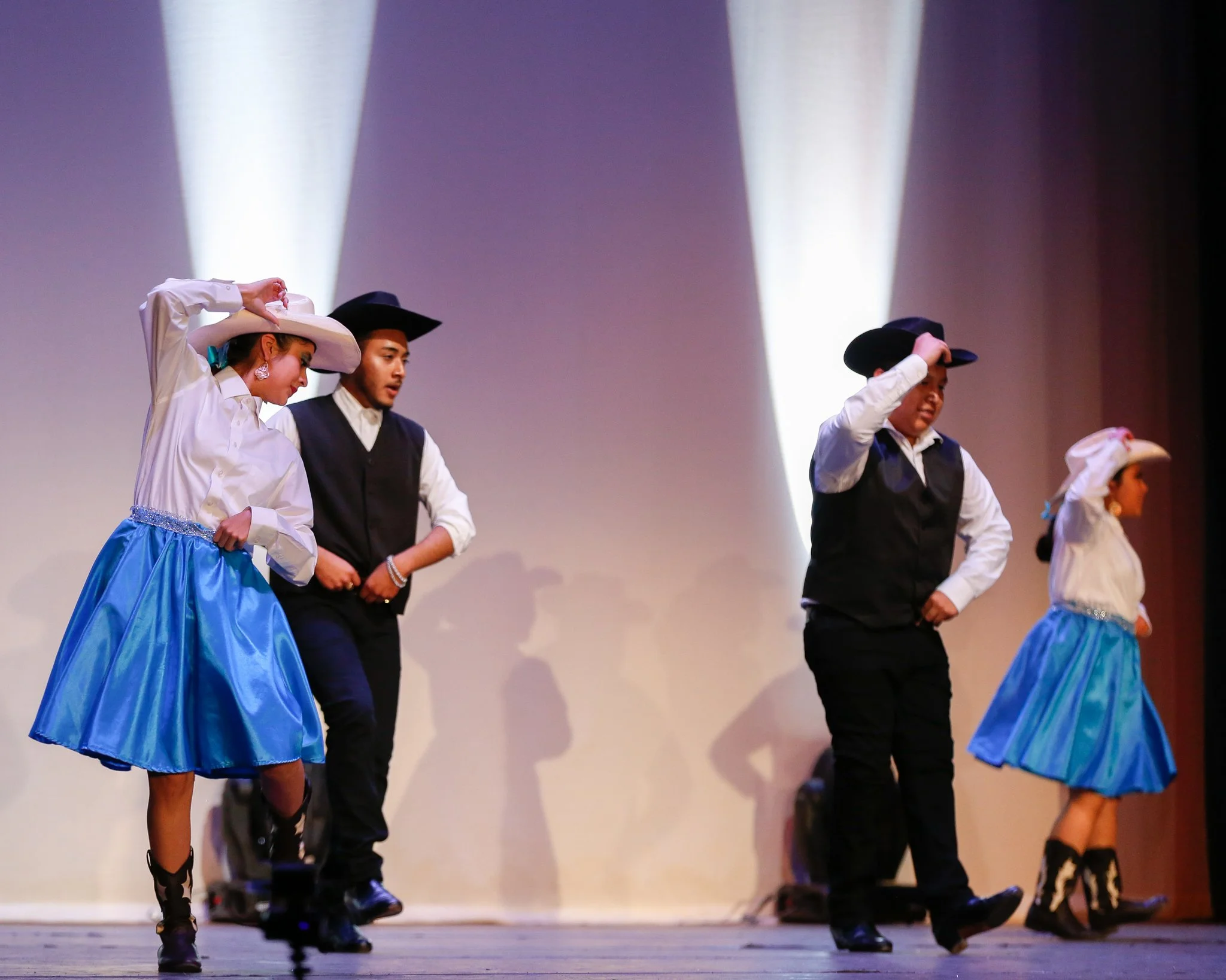 Four dancers, two women and two men, performing a traditional dance on stage, dressed in folk costumes, with the women in white blouses, blue skirts, and wide-brimmed hats, and the men in black vests, white shirts, and black hats, under stage lightin