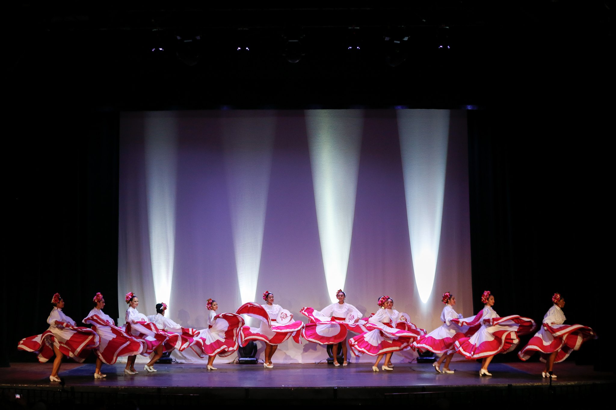 Fifteen female dancers in white blouses and pink and white skirts perform a traditional dance on a stage with purple and white lighting.