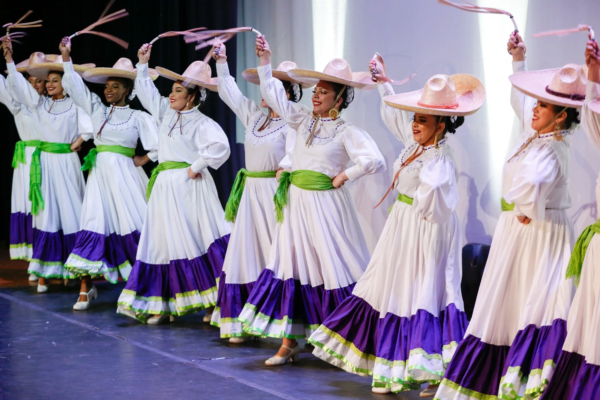 A group of women dressed in traditional Mexican folk dance costumes, with white blouses, long white skirts with purple and green accents, and large straw hats, performing a dance on stage.