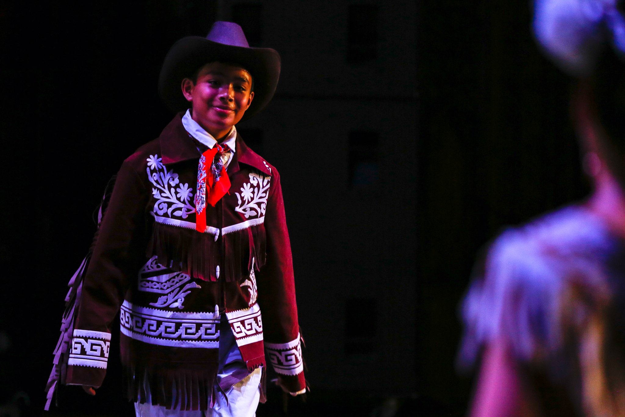 A young person dressed in traditional Mexican attire, wearing a wide-brimmed hat and a embroidered jacket, standing on a dark stage with purple and blue lighting.