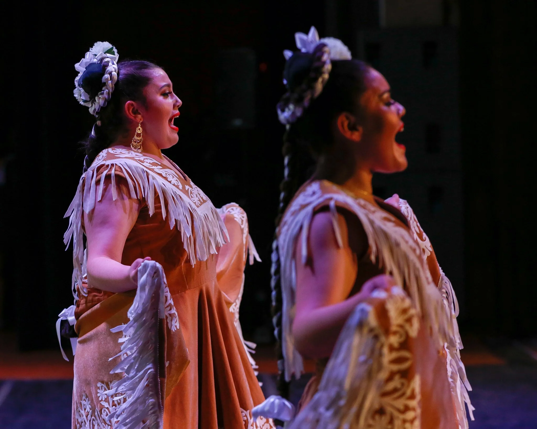 Two women dressed in traditional folkloric costumes with floral headpieces, performing a dance on stage, illuminated by stage lighting.