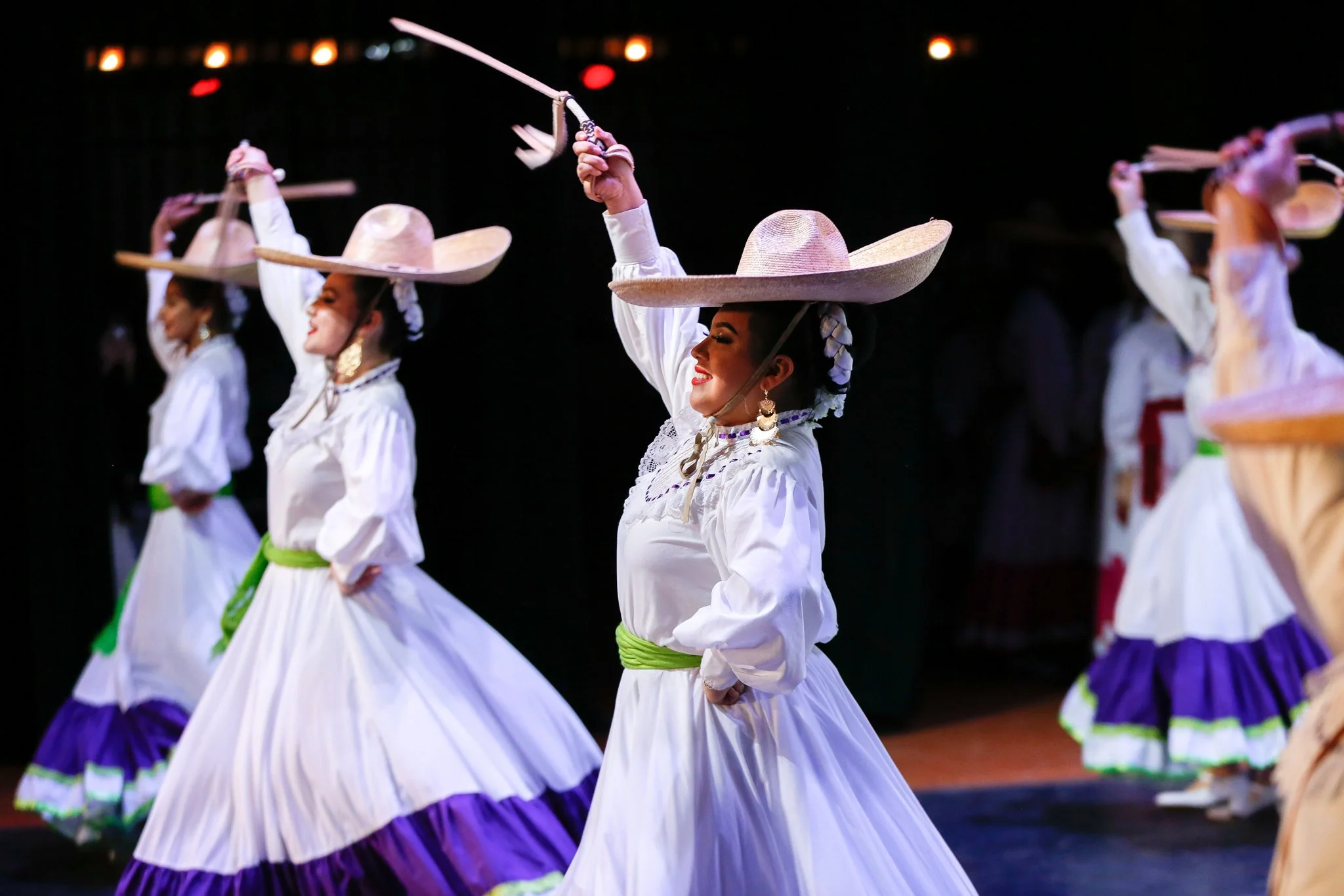 Women in traditional Mexican folkloric dresses and sombreros performing a dance with raised sticks on stage.
