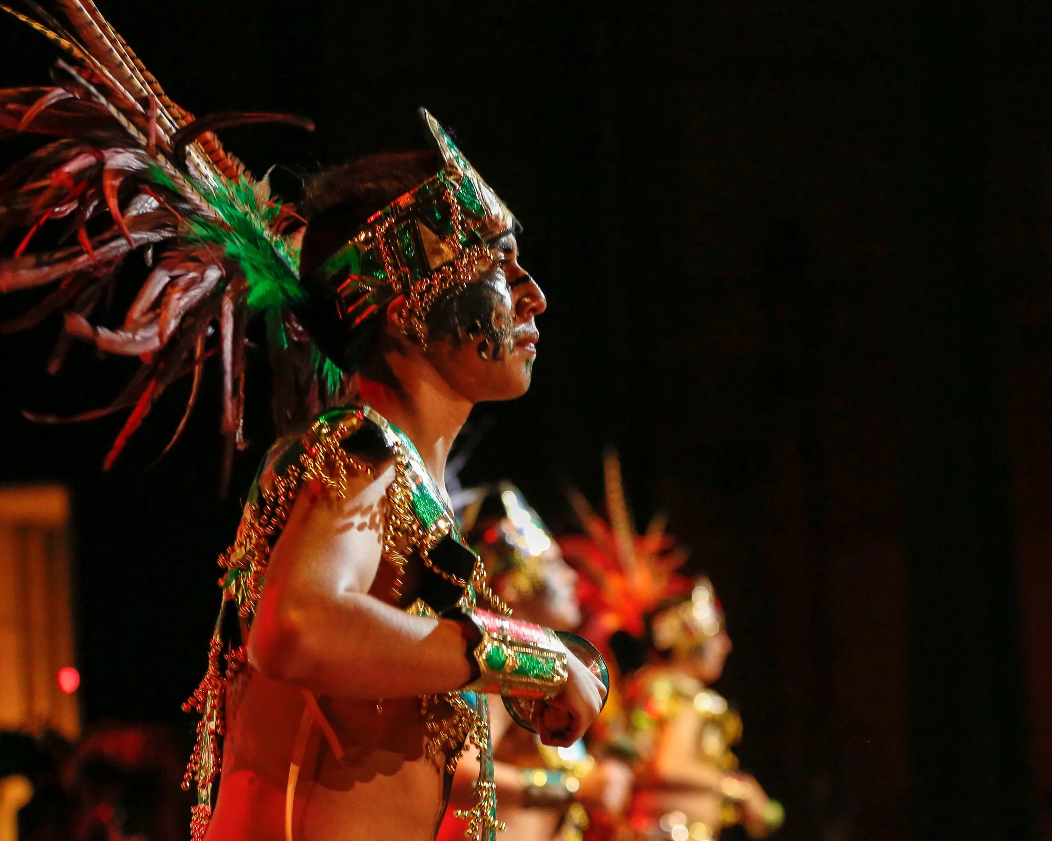 A man dressed in elaborate traditional indigenous costume with vibrant colors, feathers, and intricate decorations during a cultural ceremony at night.