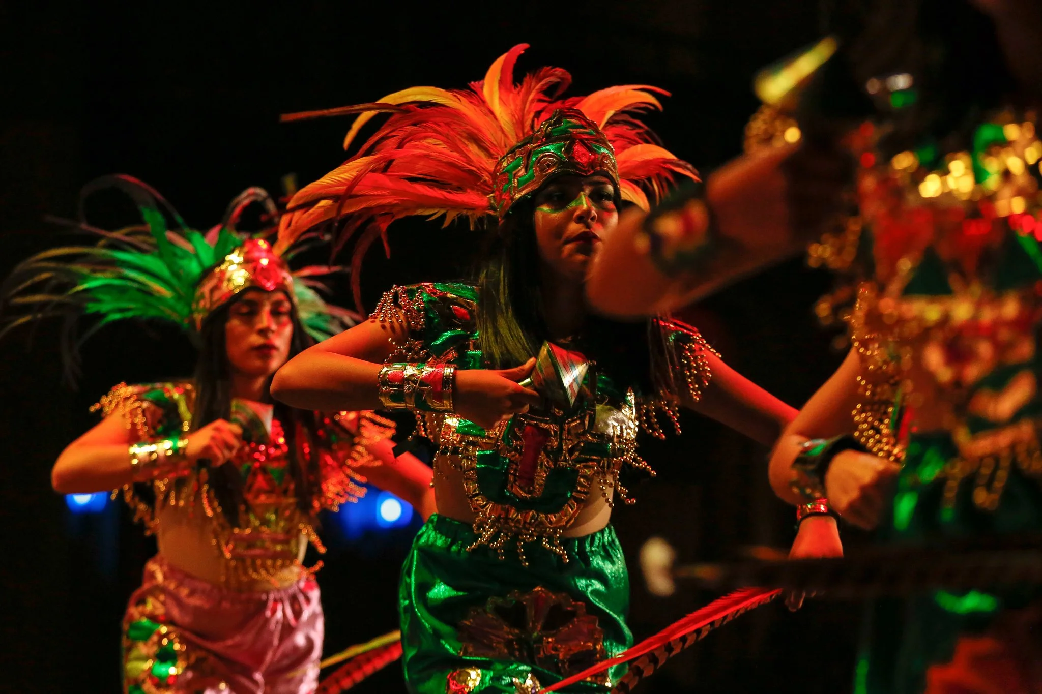 Three women in colorful, elaborate costumes with feathered headdresses dance on stage at night, in a performance with dark background and stage lighting.
