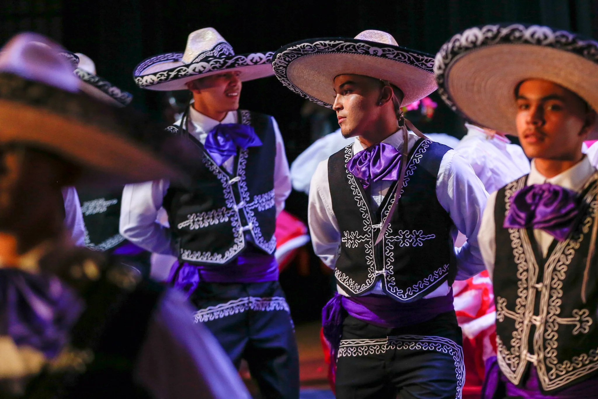 A group of young men dressed in traditional Mexican charro attire, including wide-brimmed sombreros, embroidered vests, and purple sashes, participating in a cultural dance or event.