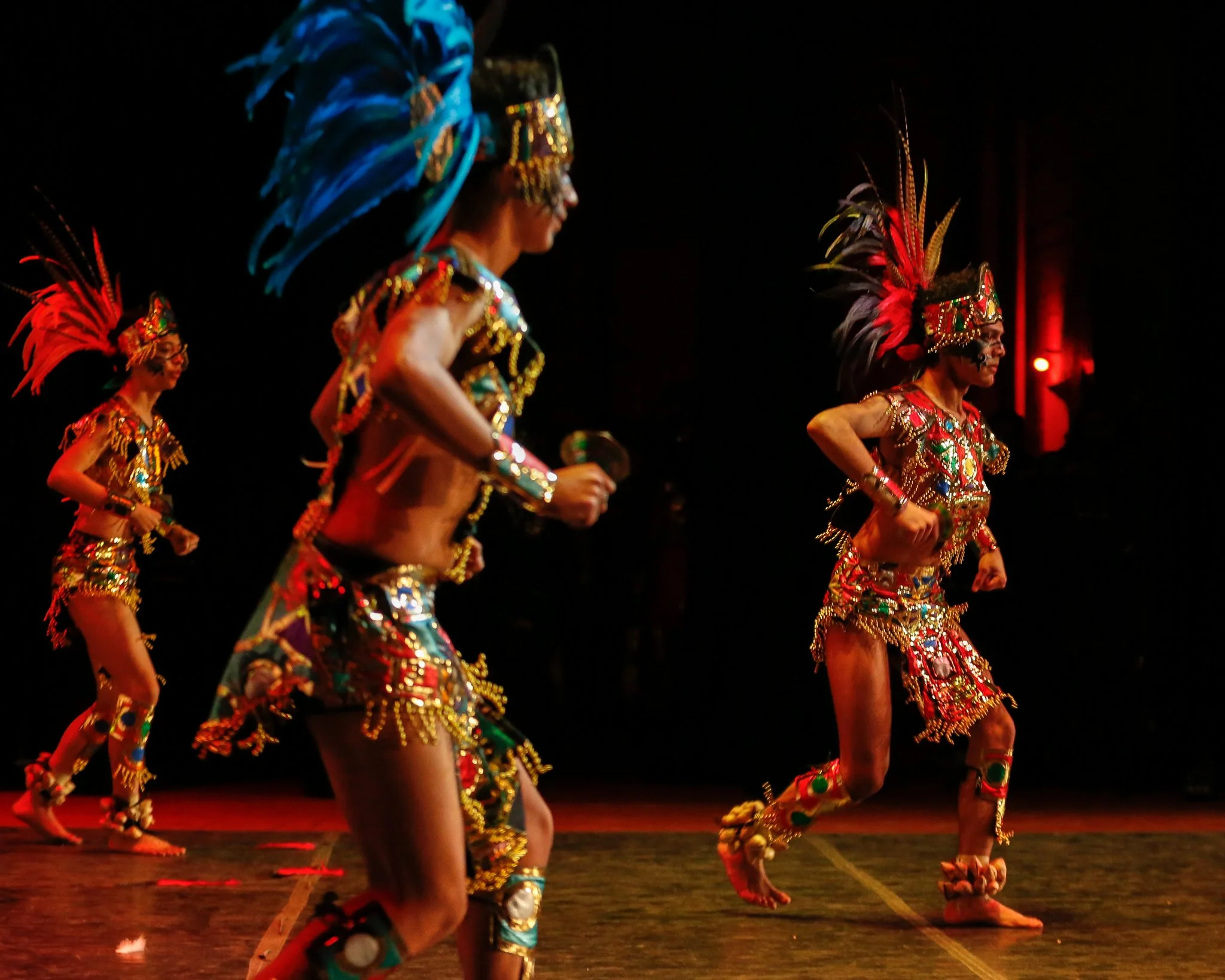 Group of dancers in vibrant, ornate traditional costumes with feathers, beads, and face paint performing on stage.