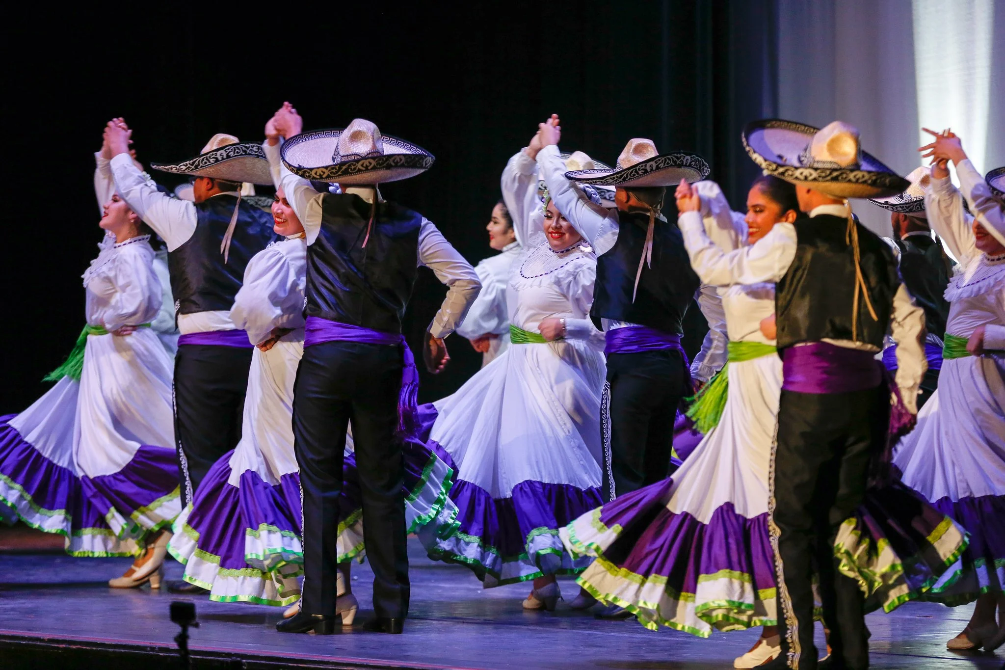 Mexican folkloric dancers performing on stage, wearing traditional colorful dresses and sombreros.