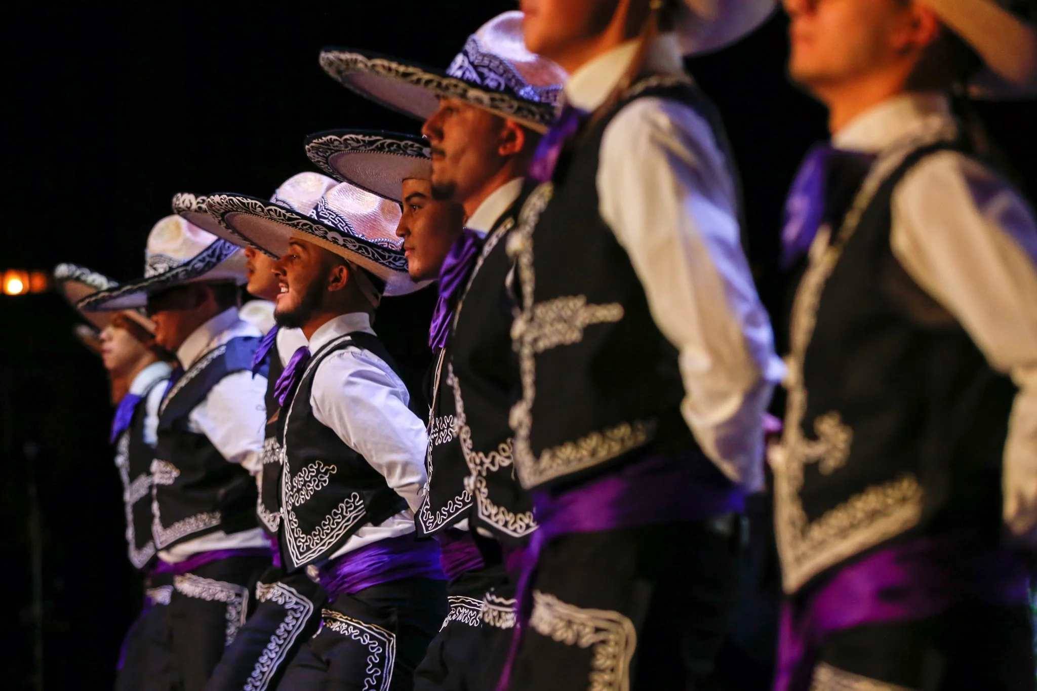 Group of Mexican dancers wearing traditional charro outfits, including sombreros, in a line formation at night.