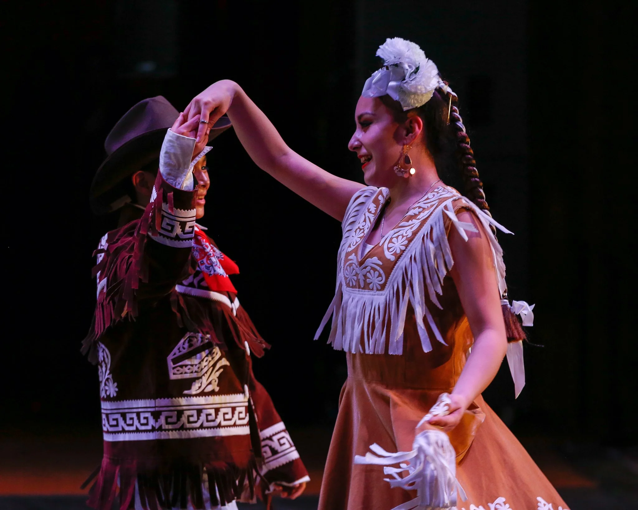 Two women in traditional Mexican folk dance attire dancing together, holding hands, with dark background