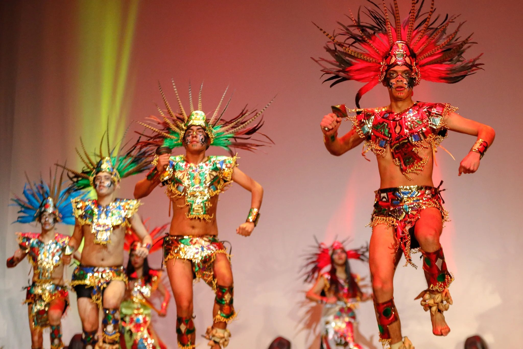 People dressed in traditional, colorful costumes with feathered headdresses performing a dance on stage.
