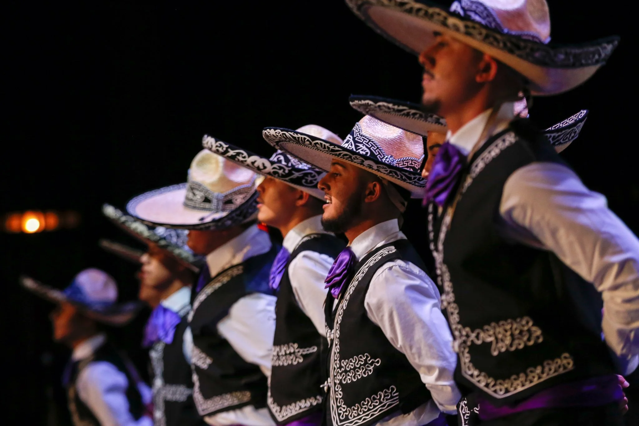 A group of people dressed in traditional Mexican attire, wearing large, decorated sombreros, and black vests with white embroidery, performing a dance at night.