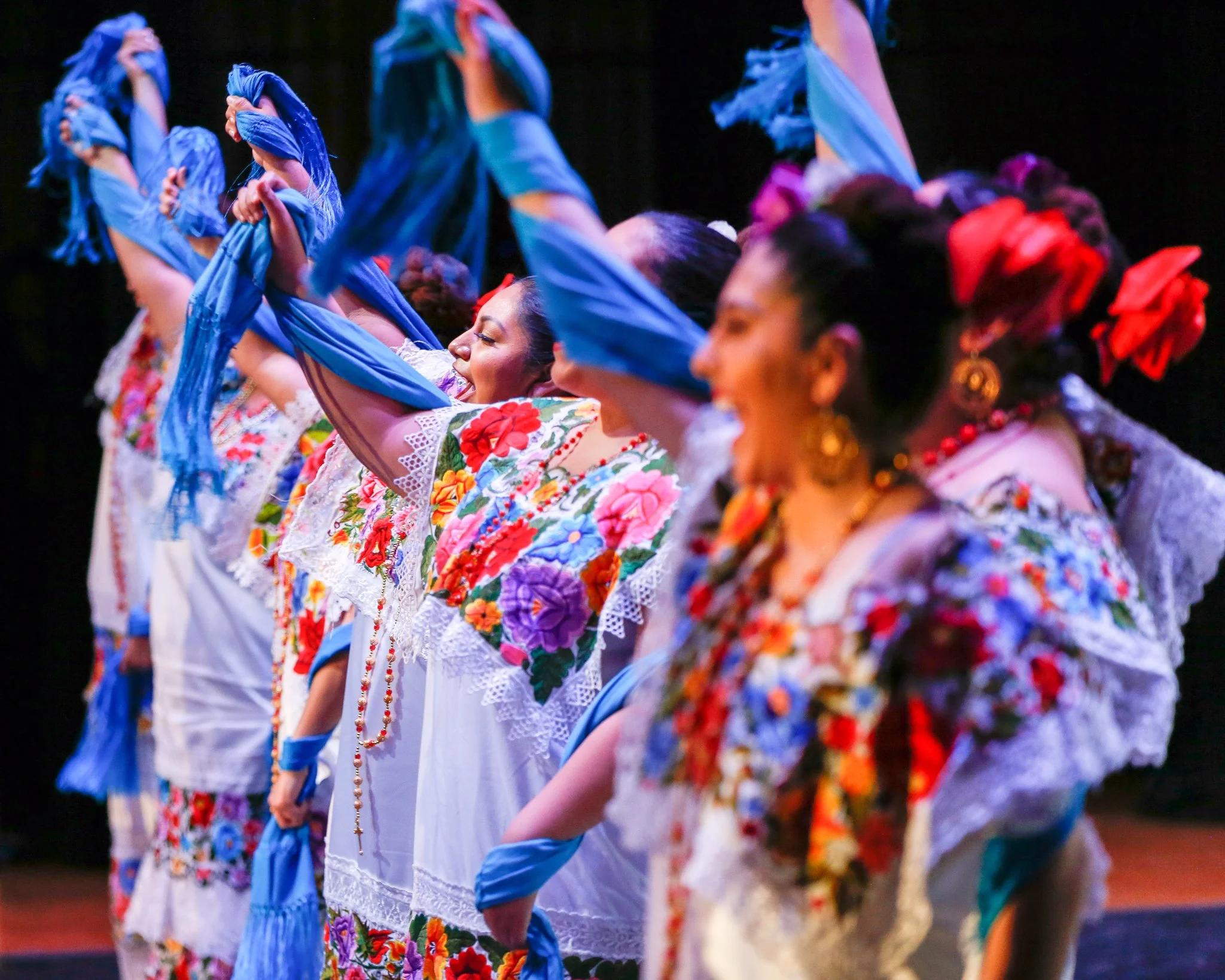 Women in traditional Mexican dresses performing a folkloric dance on stage, wearing colorful embroidered blouses, skirts, and headpieces.