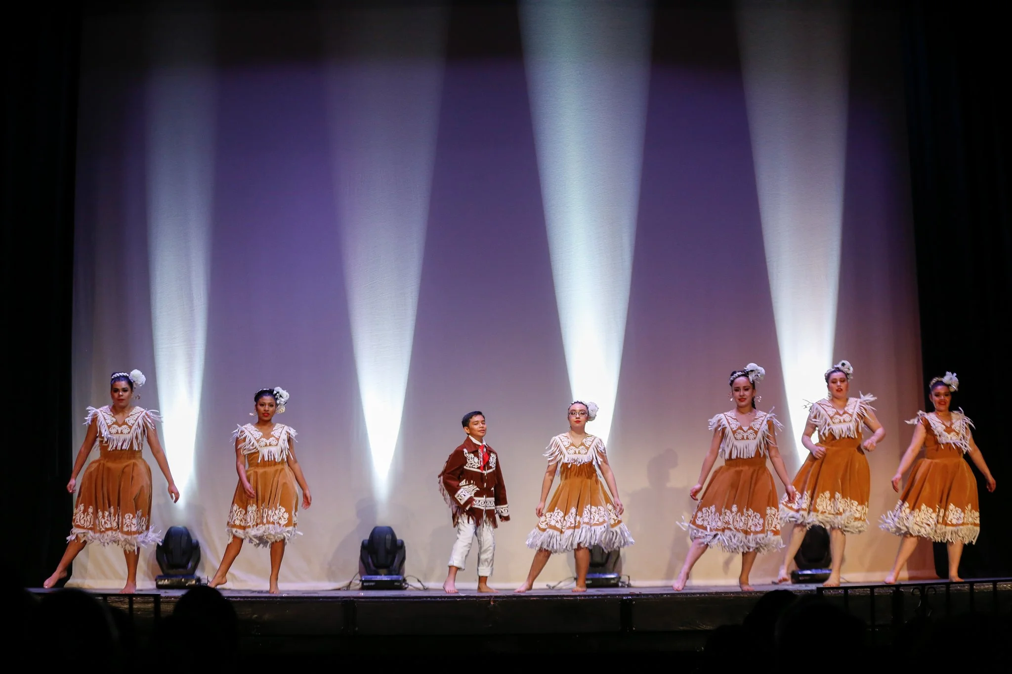 Children performing a dance on stage, dressed in traditional costumes, with female dancers in beige dresses with white lace and flowers in their hair, and a male dancer in a brown jacket with white pants, under bright stage lights.
