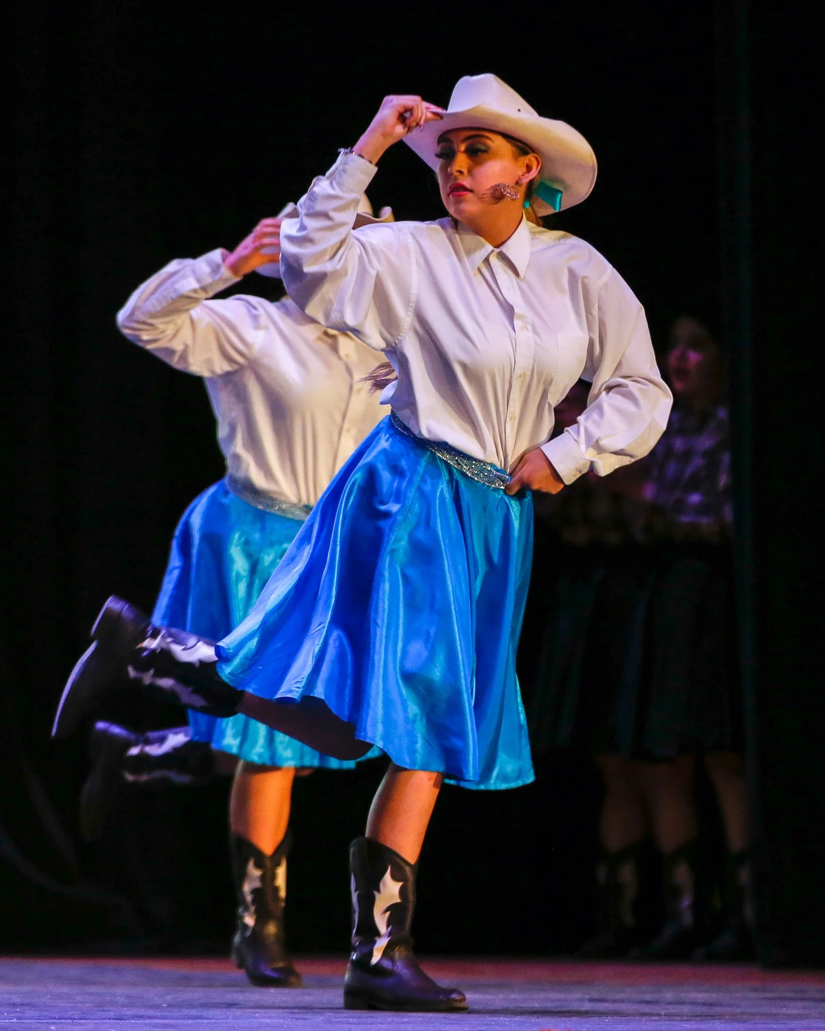 A woman dressed as a cowgirl performing a dance on stage, wearing a white cowboy hat, white shirt, blue skirt, and cowboy boots with a star design.
