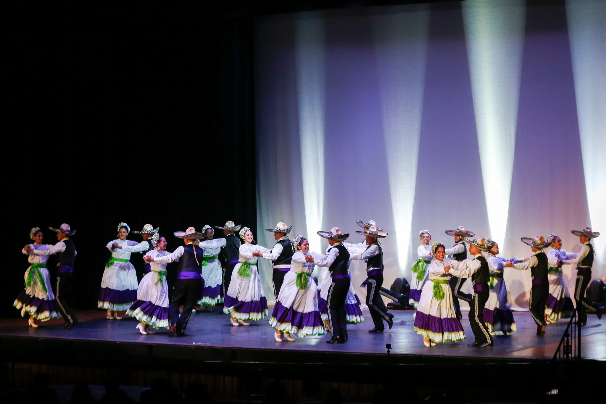 A group of dancers in traditional Mexican attire performing a folkloric dance on stage, wearing white dresses with purple and green accents and wide-brimmed sombreros, with stage lighting in the background.