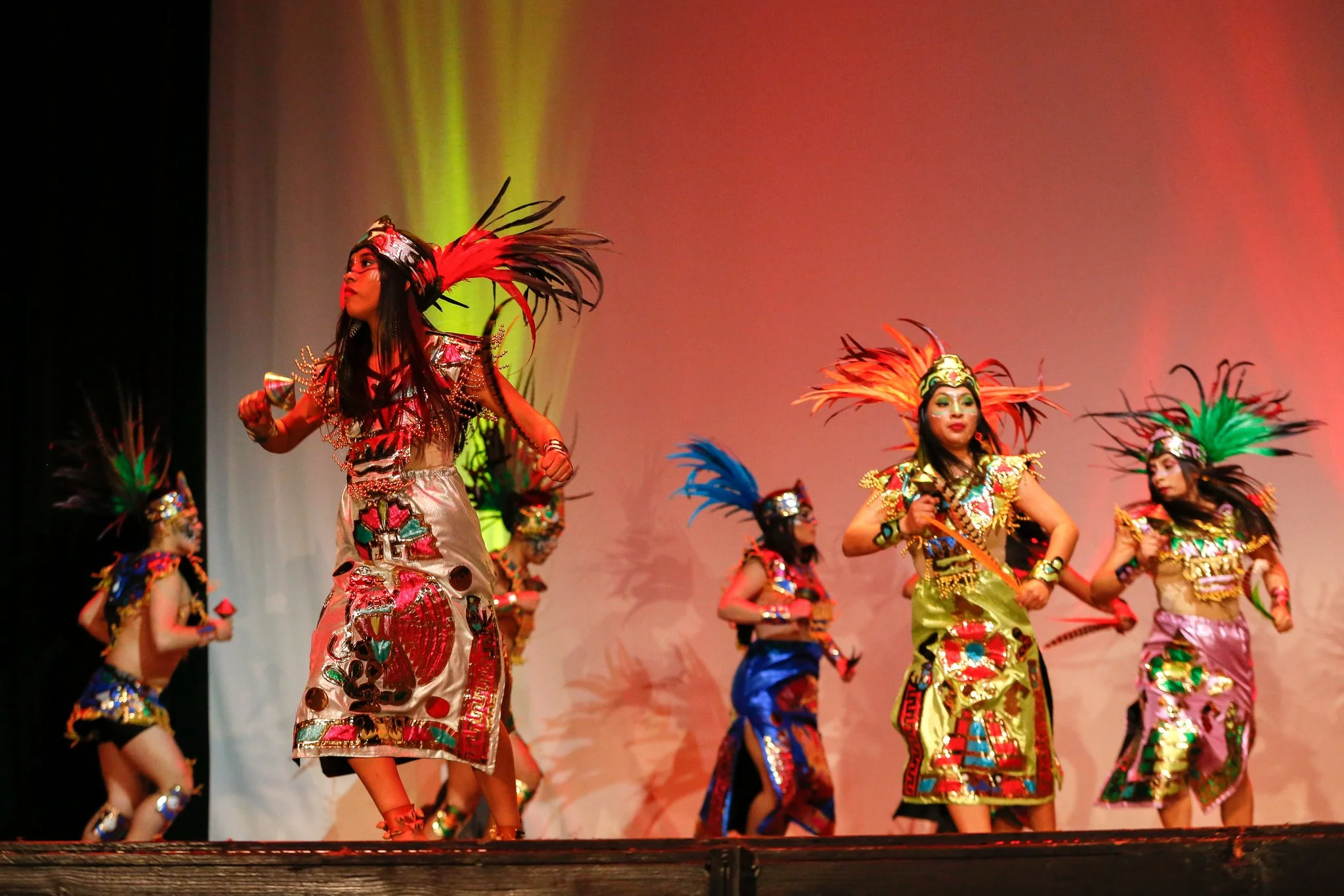 Group of performers in colorful traditional costumes with feathered headdresses on stage, dancing.