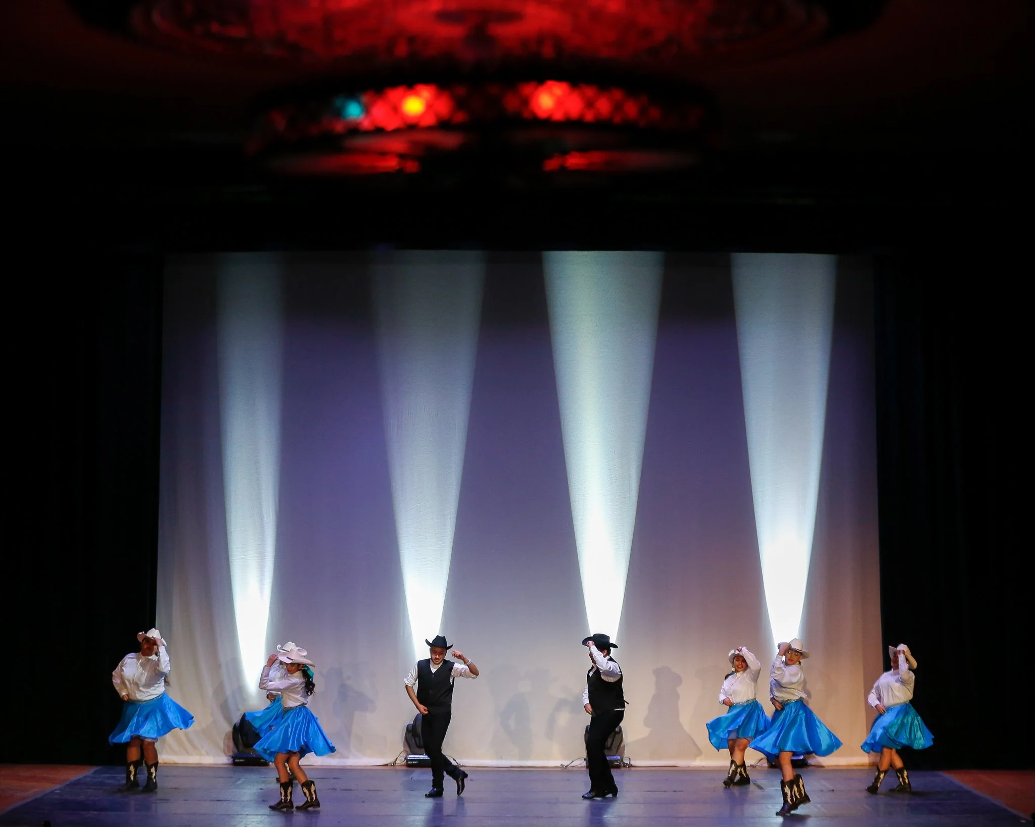 Group of six dancers performing on stage with blue skirts, white tops, and cowboy hats, illuminated by stage lights and a backdrop.