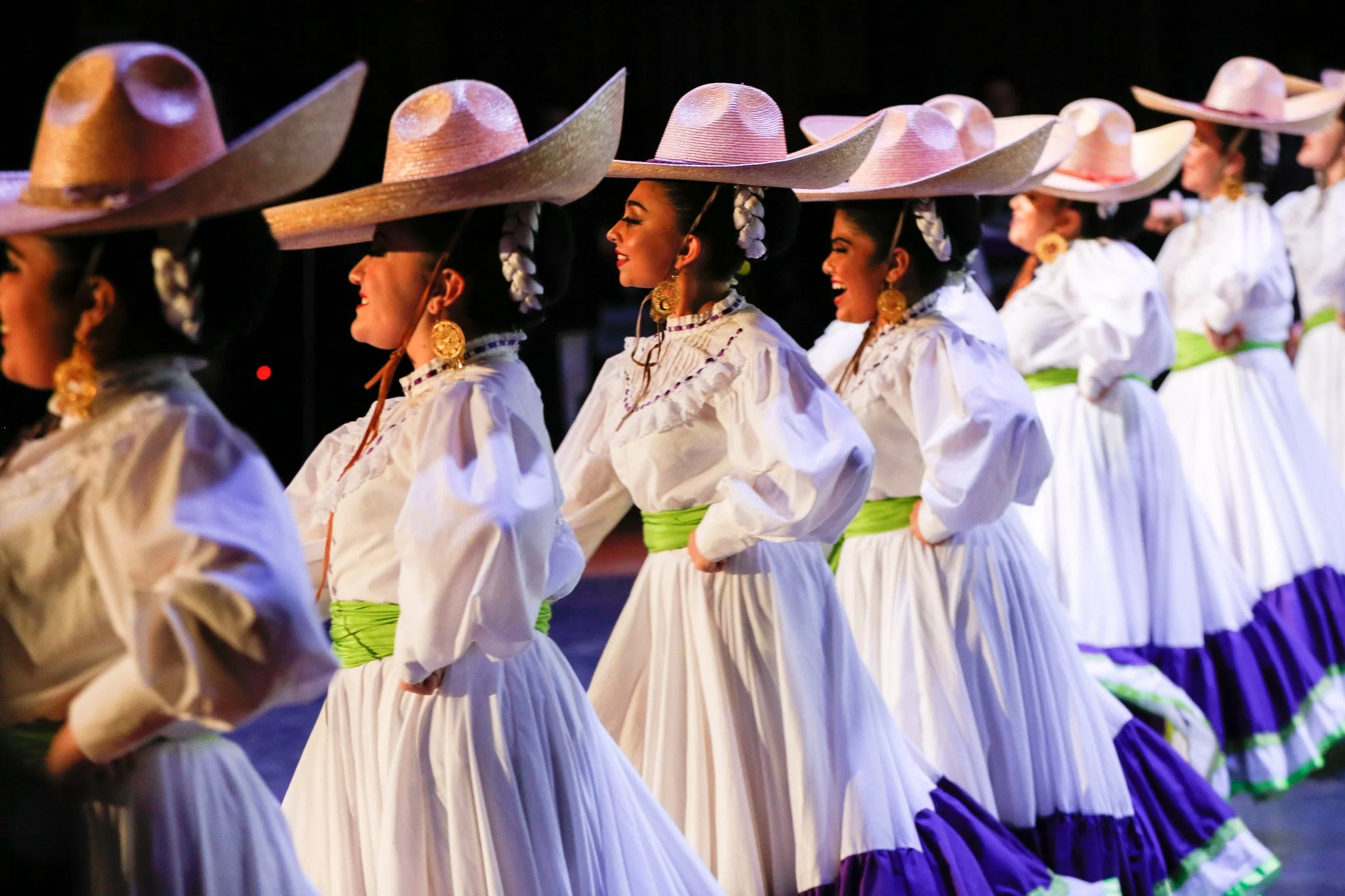 Mexican folkloric dancers in traditional white dresses with green and purple accents and large sombrero hats performing a cultural dance.