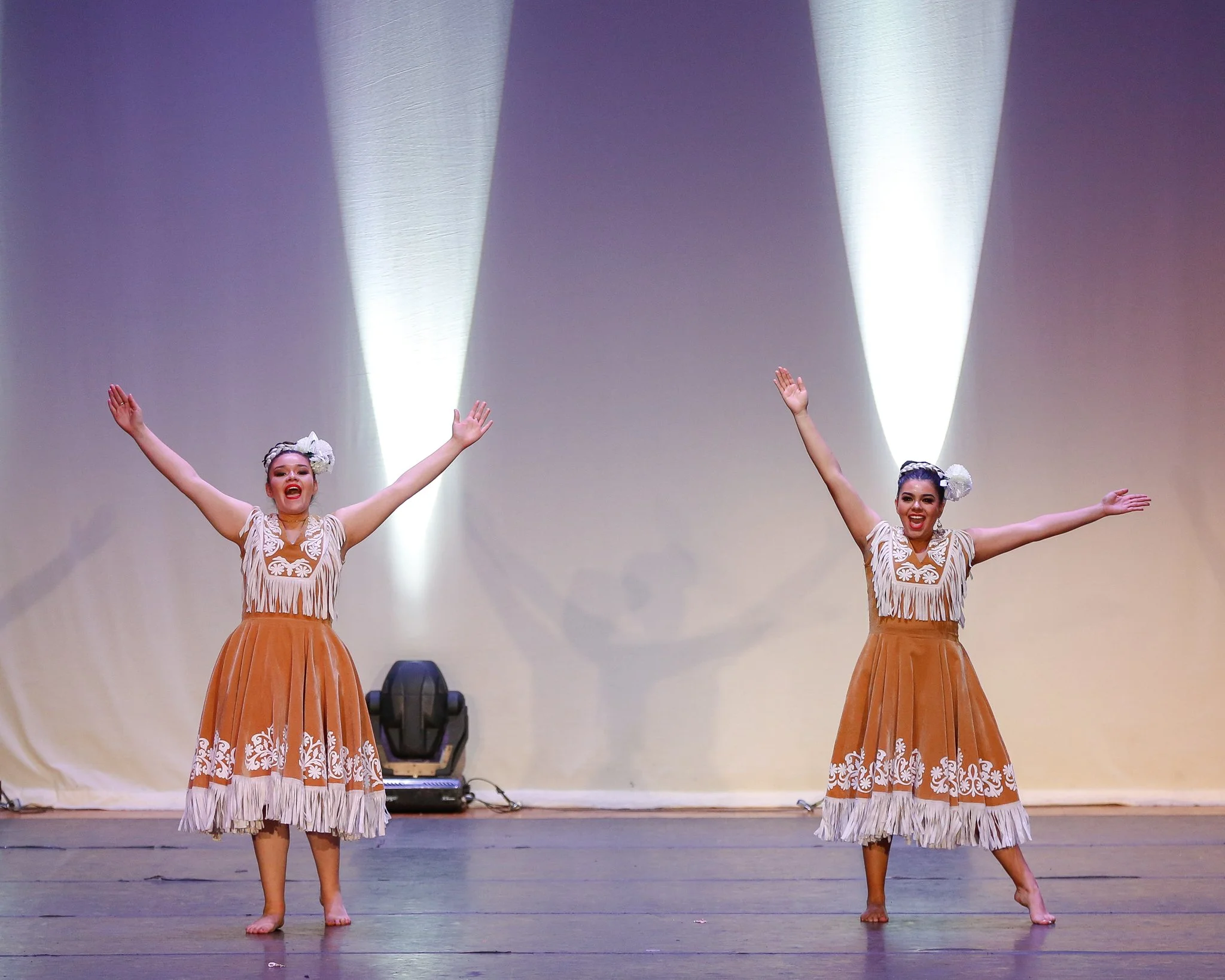 Two women in traditional Polynesian costumes performing a dance on stage with beige background and bright white spotlights.