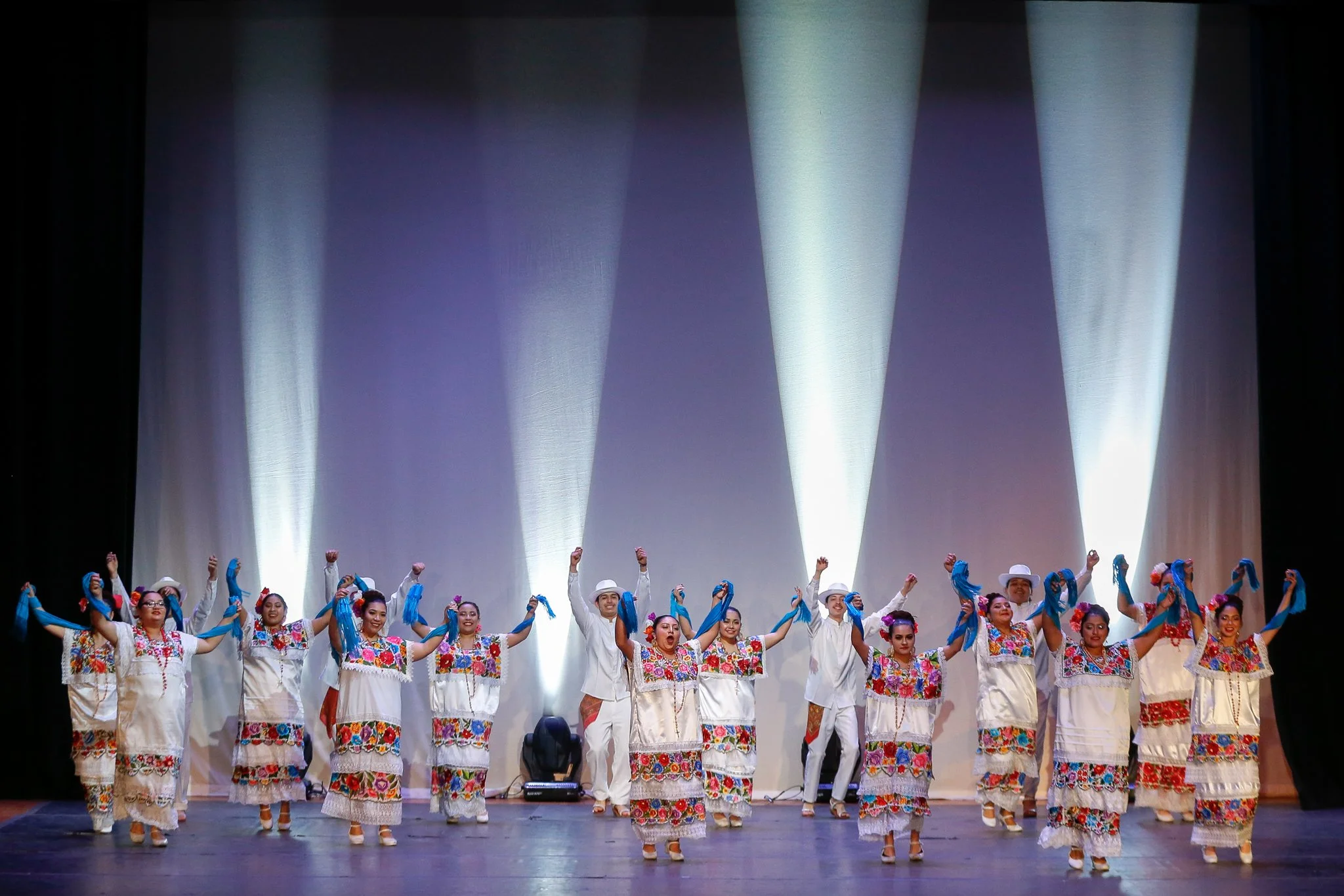 Group of performers on stage in traditional Mexican attire, holding blue scarves, dancing during a cultural performance.