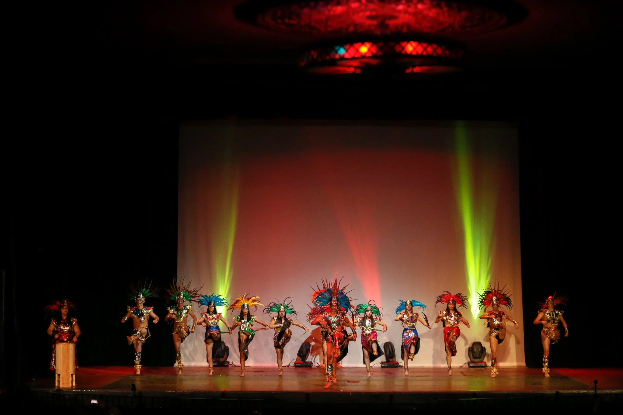 A group of performers on stage dressed in colorful costumes with feathers, dancing in a choreographed routine with stage lights and a plain backdrop.