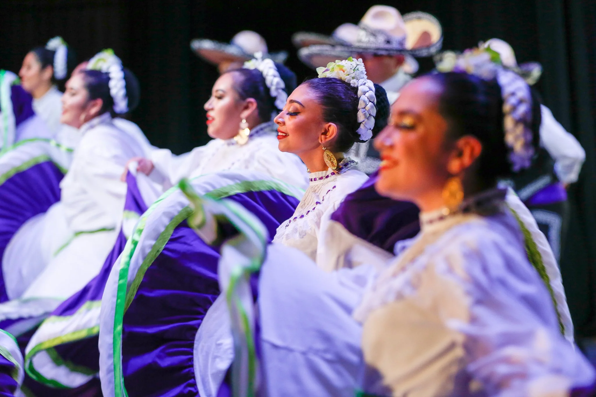 Women in traditional Mexican dresses performing a folkloric dance, with white, purple, and green elaborate costumes and floral hair accessories.