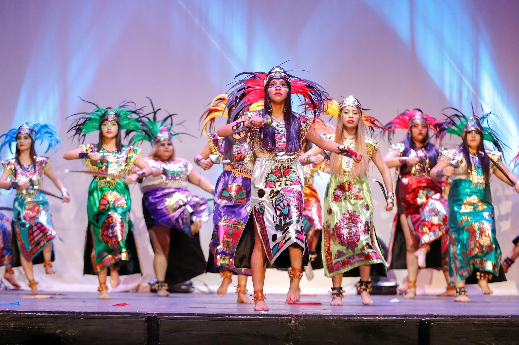 Group of young girls performing a traditional dance on stage, dressed in colorful tribal costumes with feathers and beads.