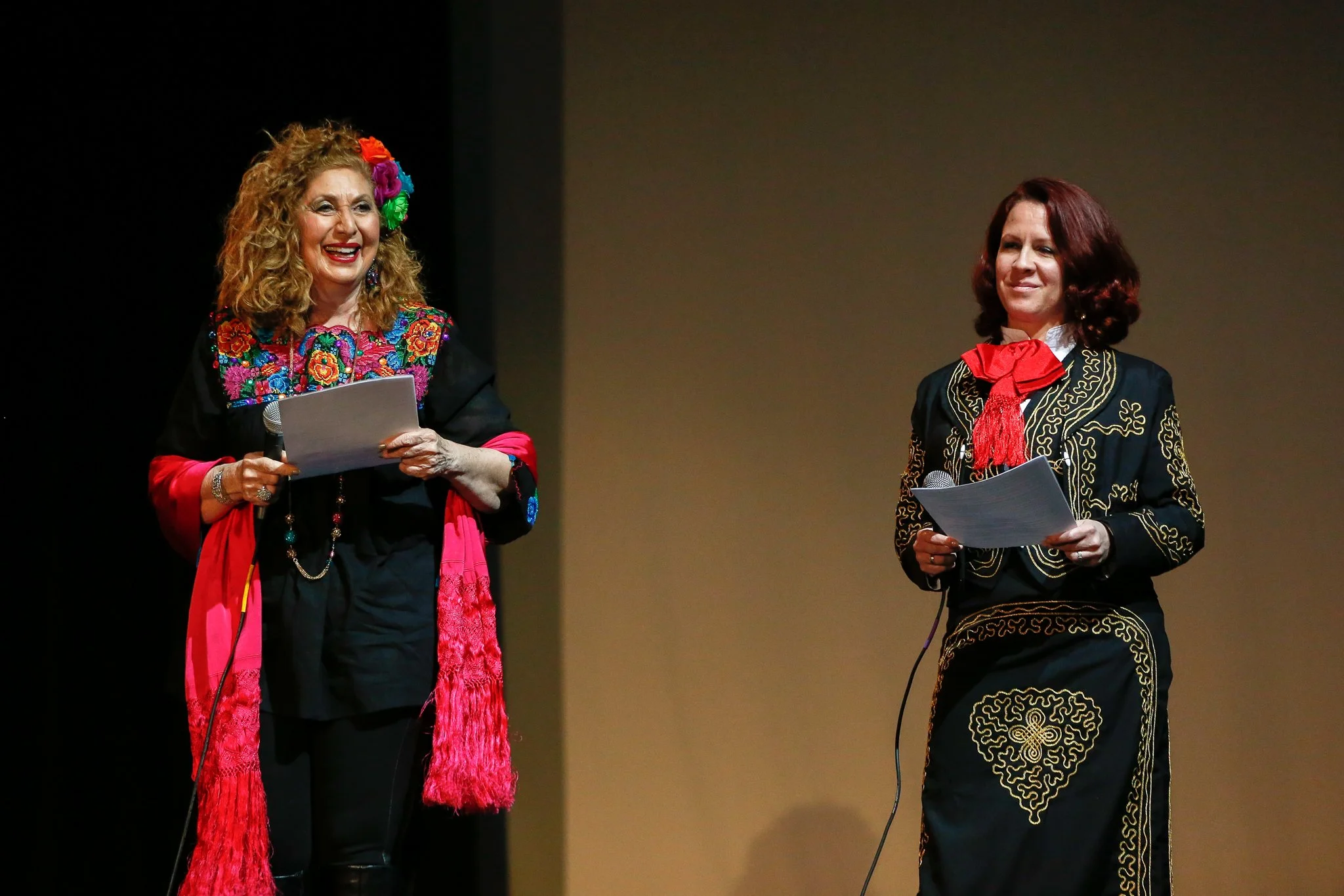 Two women on stage dressed in traditional Mexican attire, holding papers, one smiling and the other with a neutral expression.
