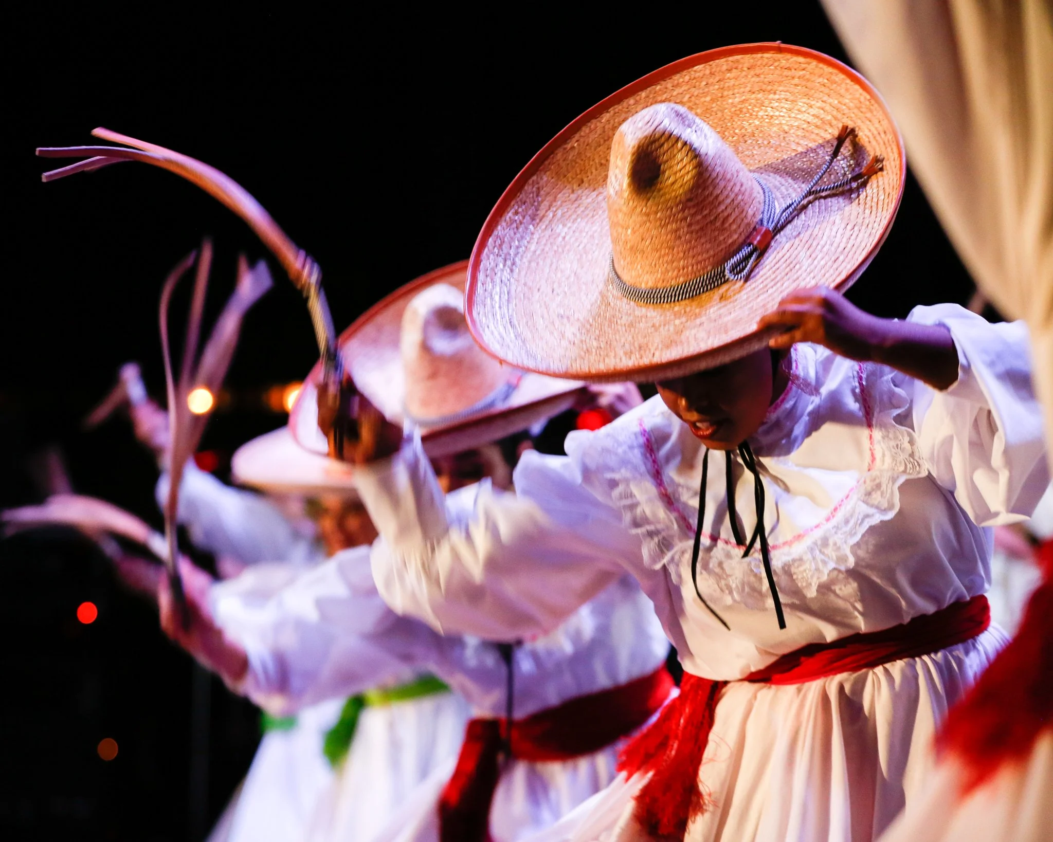 Women dressed in traditional white Mexican dresses with red sashes perform a folkloric dance outdoors at night, wearing large straw sombreros.