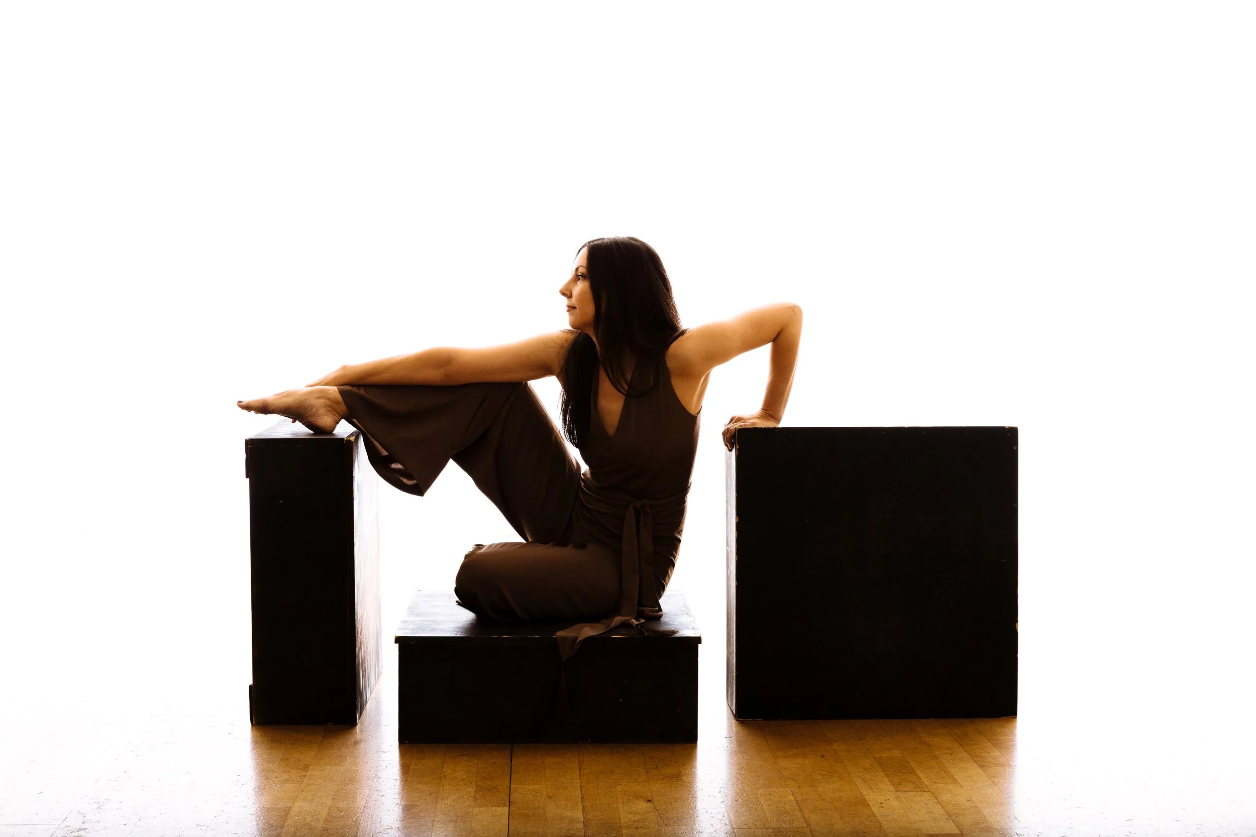 A woman in a dark dress sitting on a black box, extending one leg onto another box and leaning back with her arm supporting her.