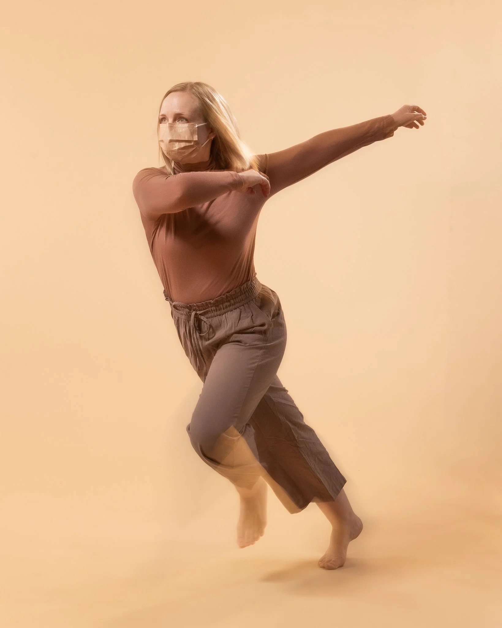 A woman wearing a face mask performing a dance pose against a beige background.