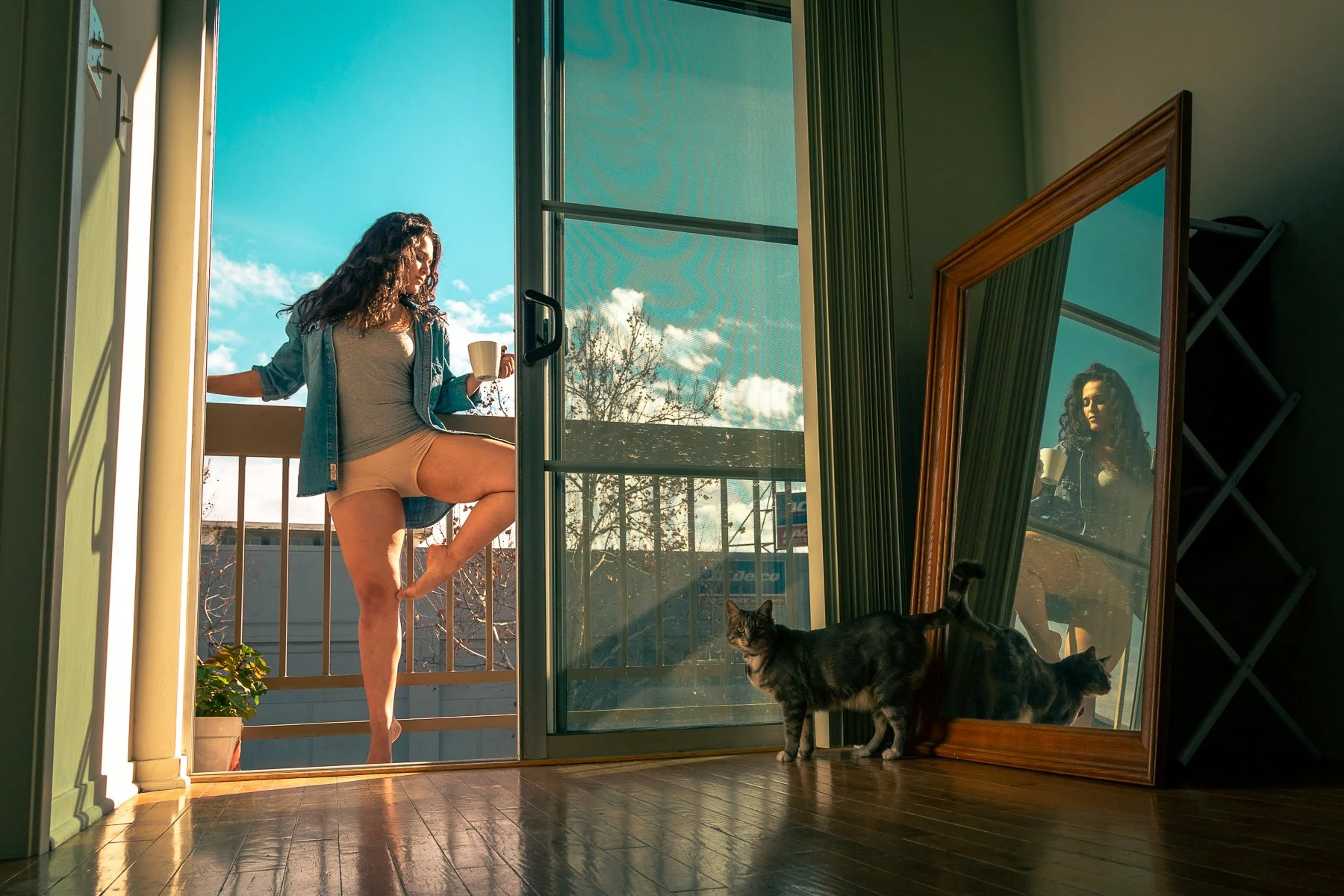 A woman standing on a balcony, holding a mug, seen from inside through an open sliding glass door. Two cats, one near the door and another reflected in a leaning mirror, are also inside the room. The sky is partly cloudy, and the scene is bathed in w