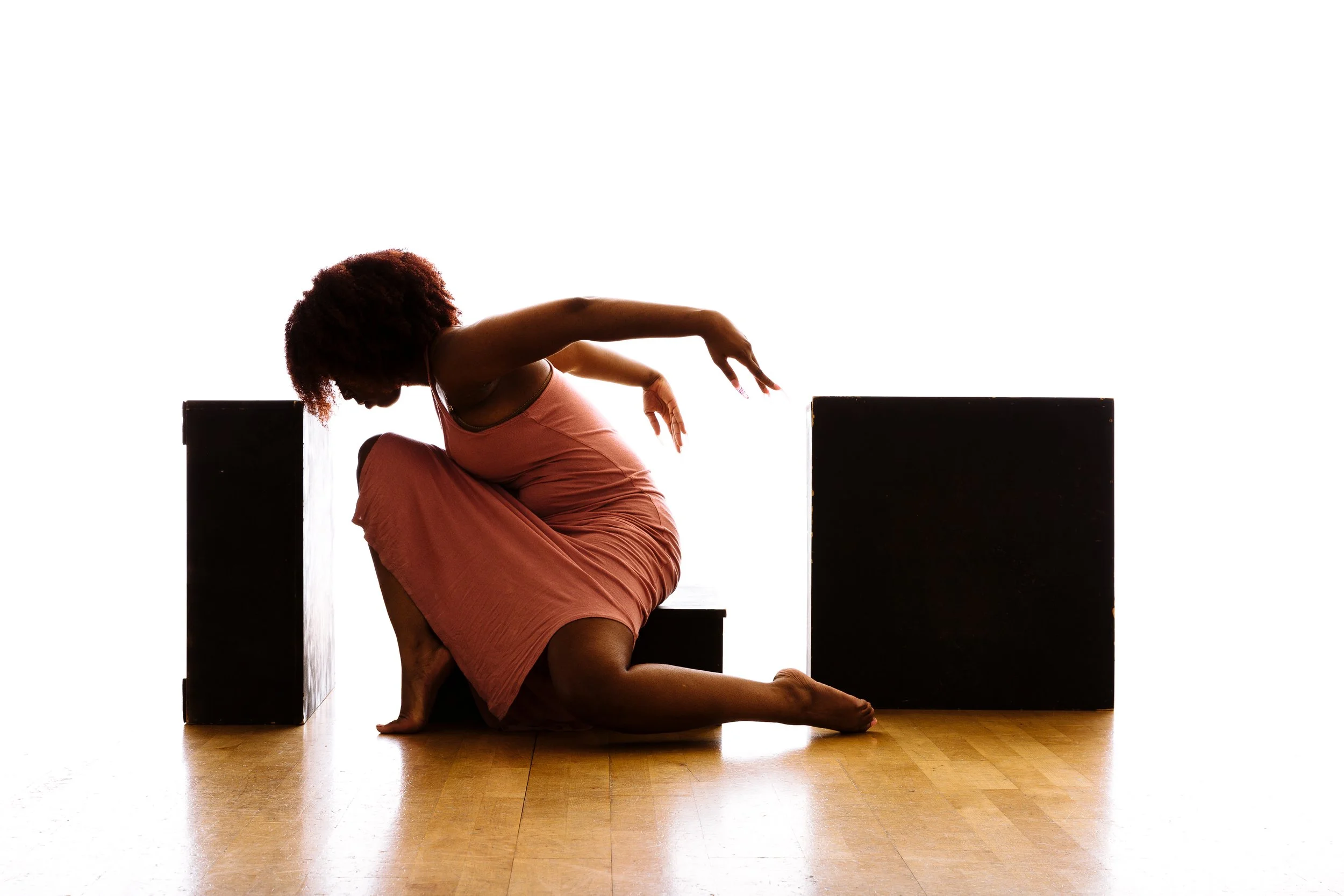 A woman with curly hair and a pink dress kneels on a wooden floor, balancing with arms extended and head lowered between two black rectangular blocks, against a plain white background.