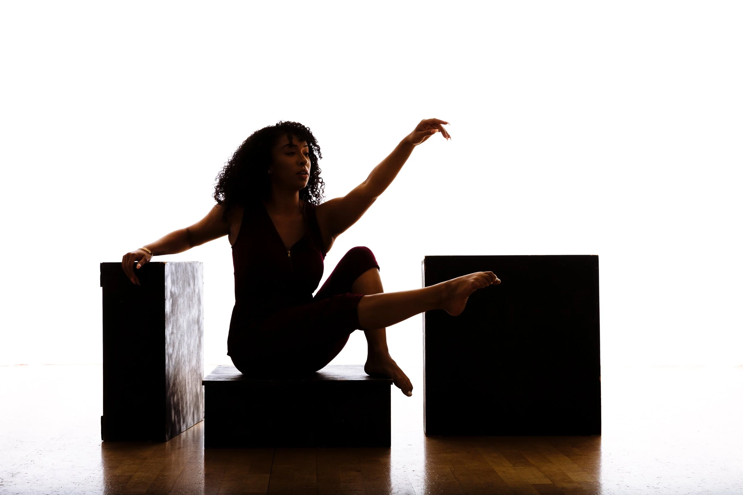 A silhouette of a woman with curly hair sitting on a black platform, reaching out with one arm, with black boxes on either side of her, against a white background.
