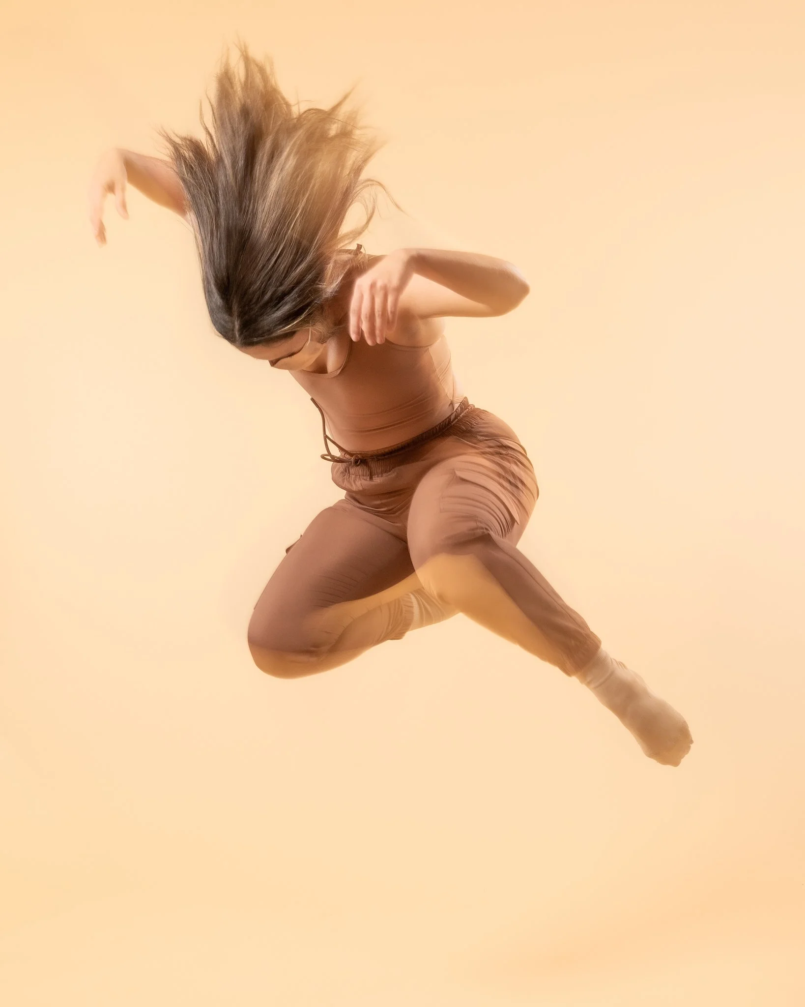 A woman in brown athletic clothes is captured mid-air against a beige background. She has long, flowing hair and is in a dynamic pose, possibly dancing or jumping.