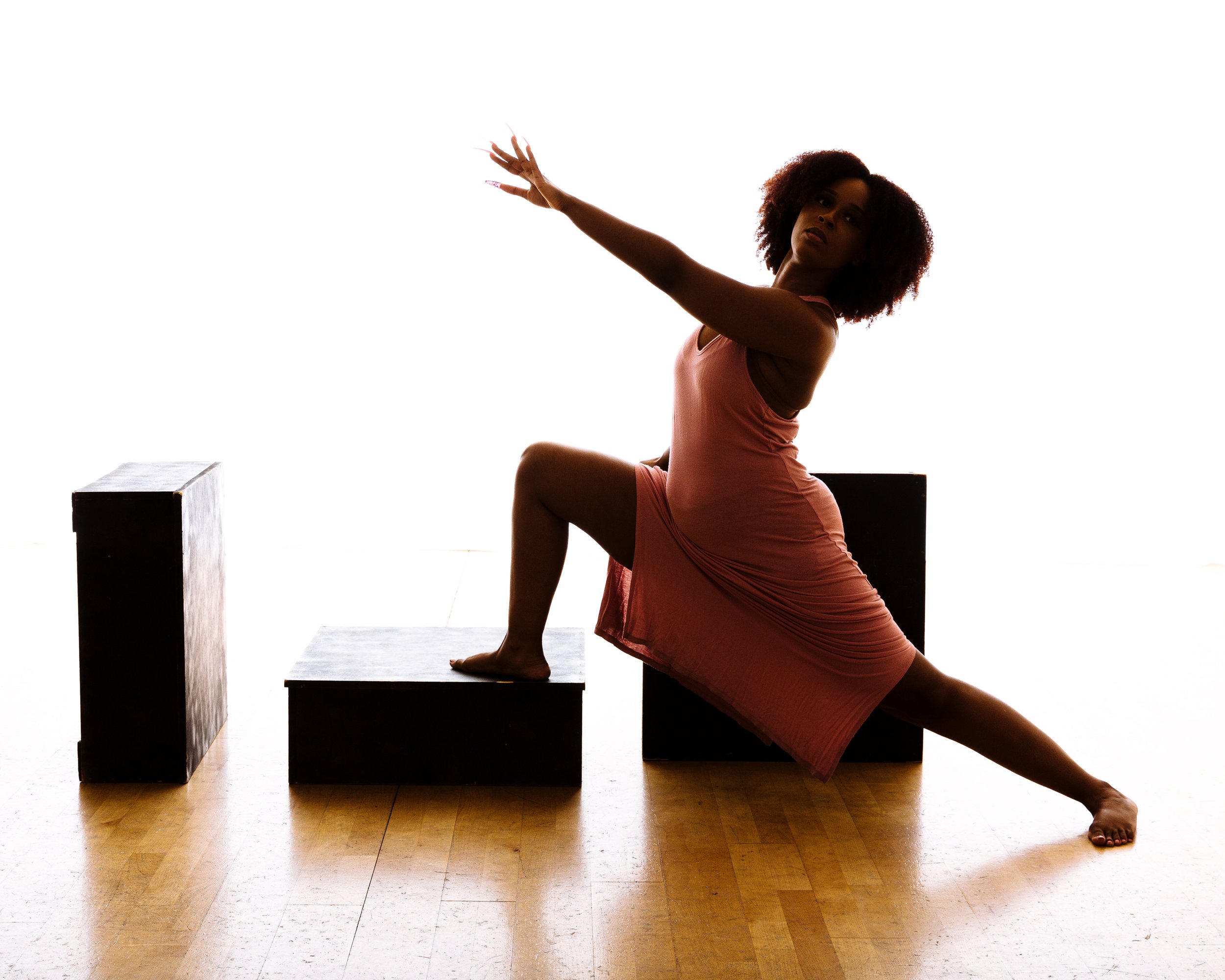A woman in a pink dress performing a dance pose with her left hand reaching forward, her right hand extended upward, one foot on a low black cube, and the other leg stretched out behind her, against a bright white background.