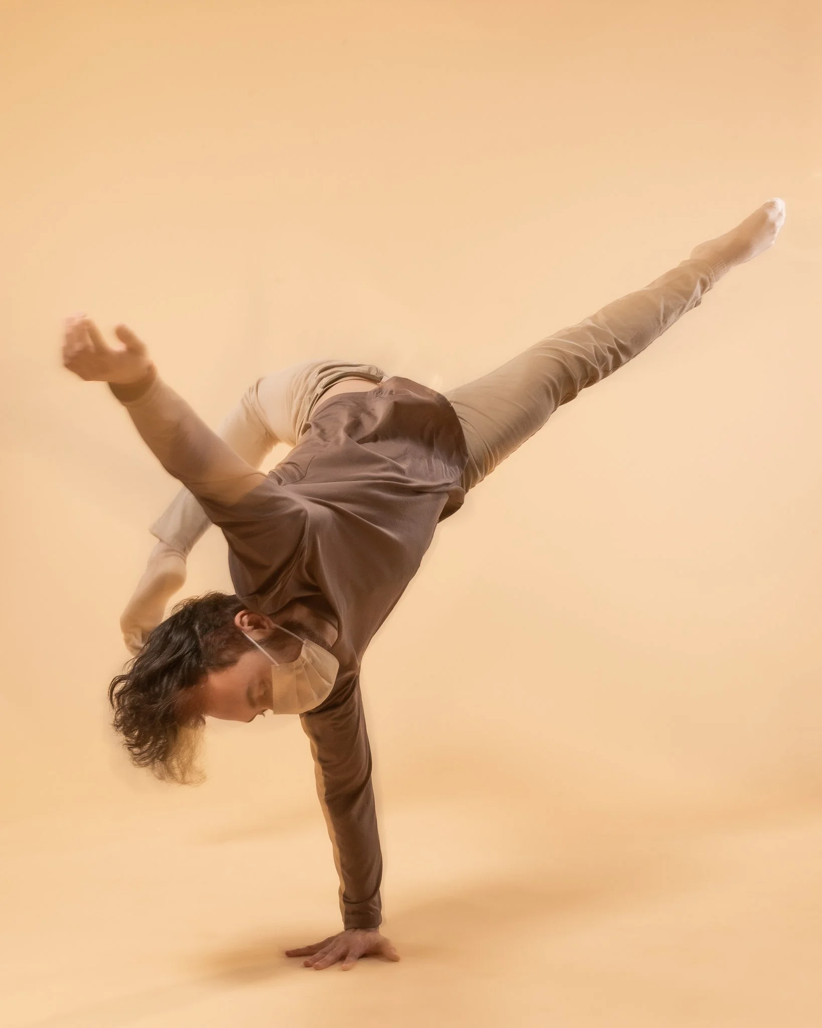 Person performing a handstand dance move with one hand on the ground, wearing a face mask, beige pants, a brown long-sleeve shirt, and a beige top against a neutral background.