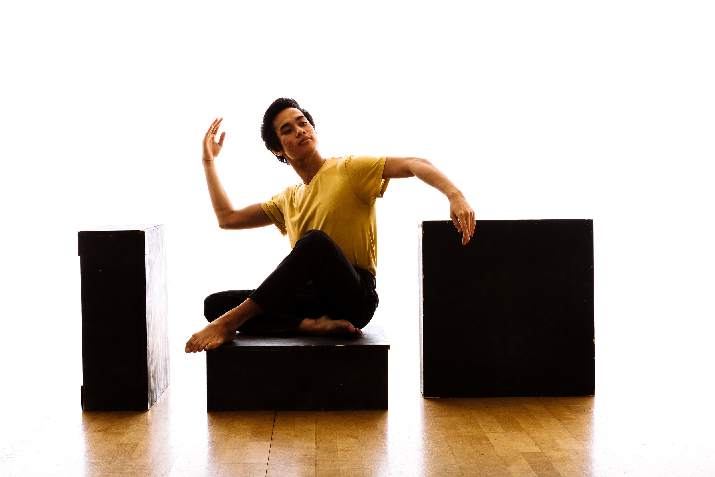 A person with short dark hair wearing a yellow shirt and black pants sitting cross-legged on a black platform, with two black cubes on either side, one taller and one shorter, against a white background.