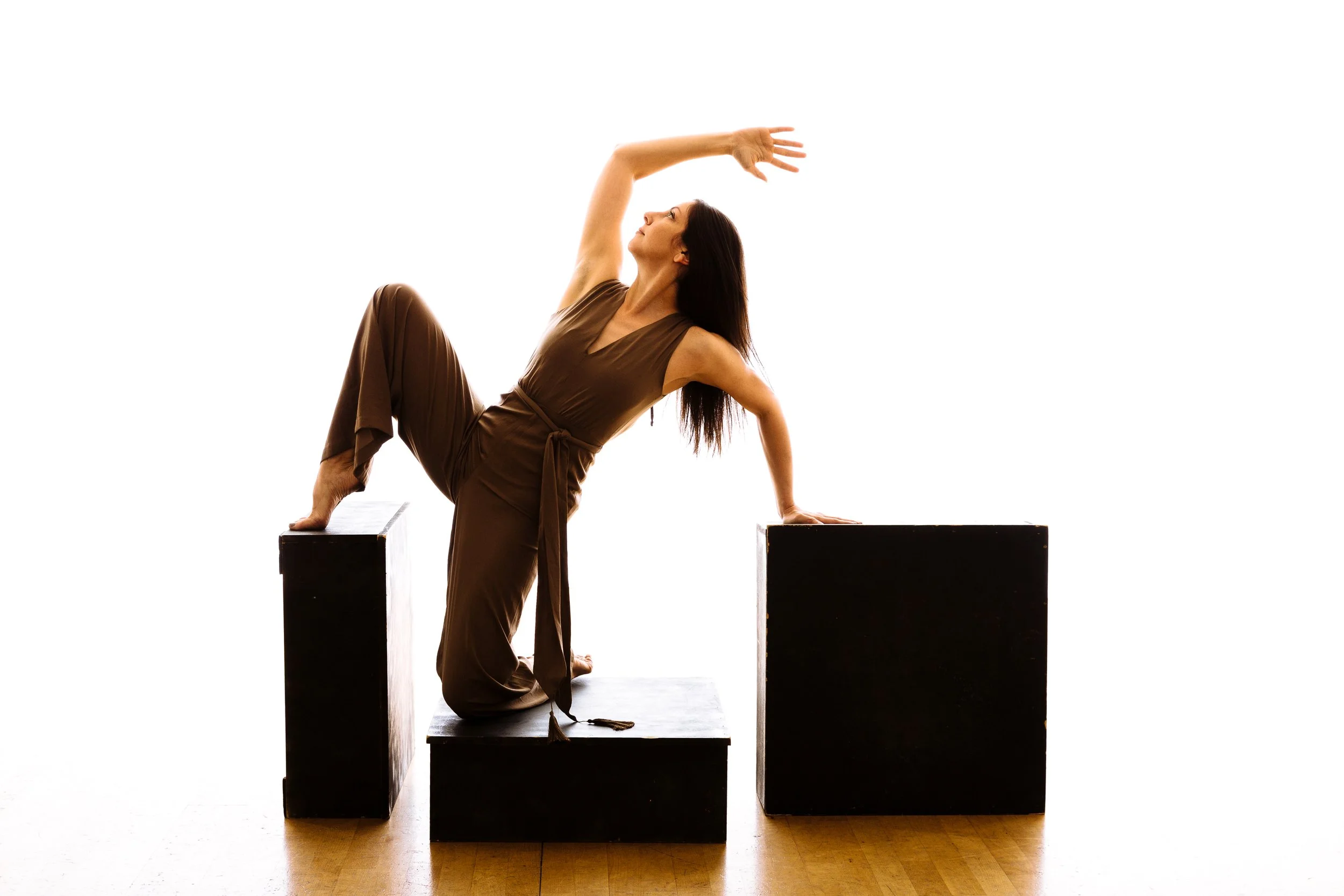 A woman in a brown jumpsuit balancing on black rectangular blocks, striking a dance pose with one hand on the block and the other raised above her head against a white background.